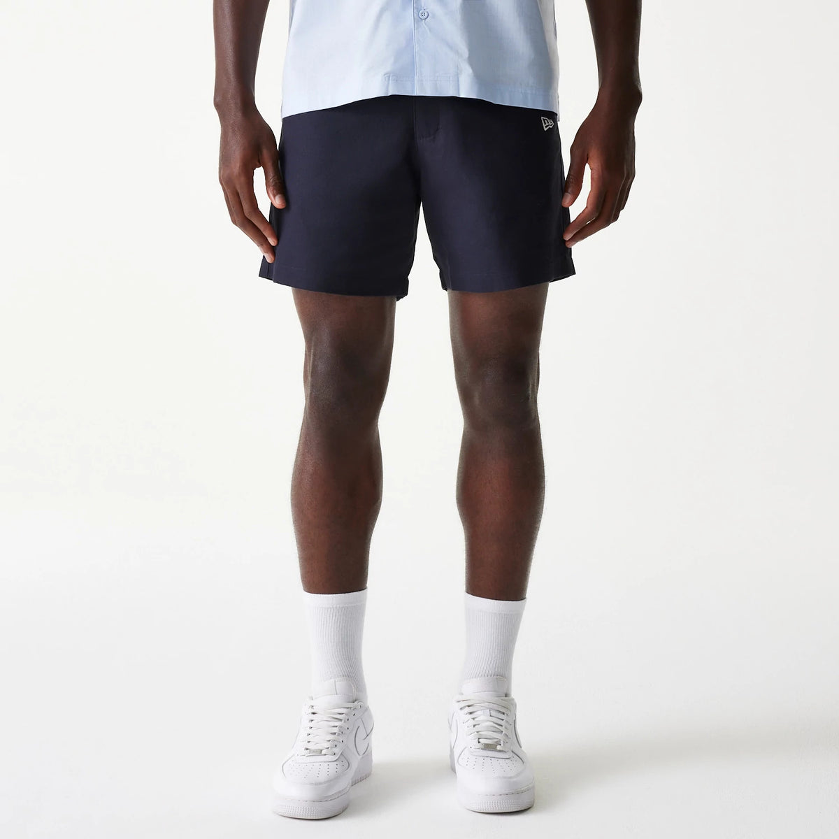 New Era Navy Chino Shorts, front view showing the navy fabric with a debossed New Era logo on the left leg, the shorts cut above the knee paired with white sneakers and socks, creating a casual look.