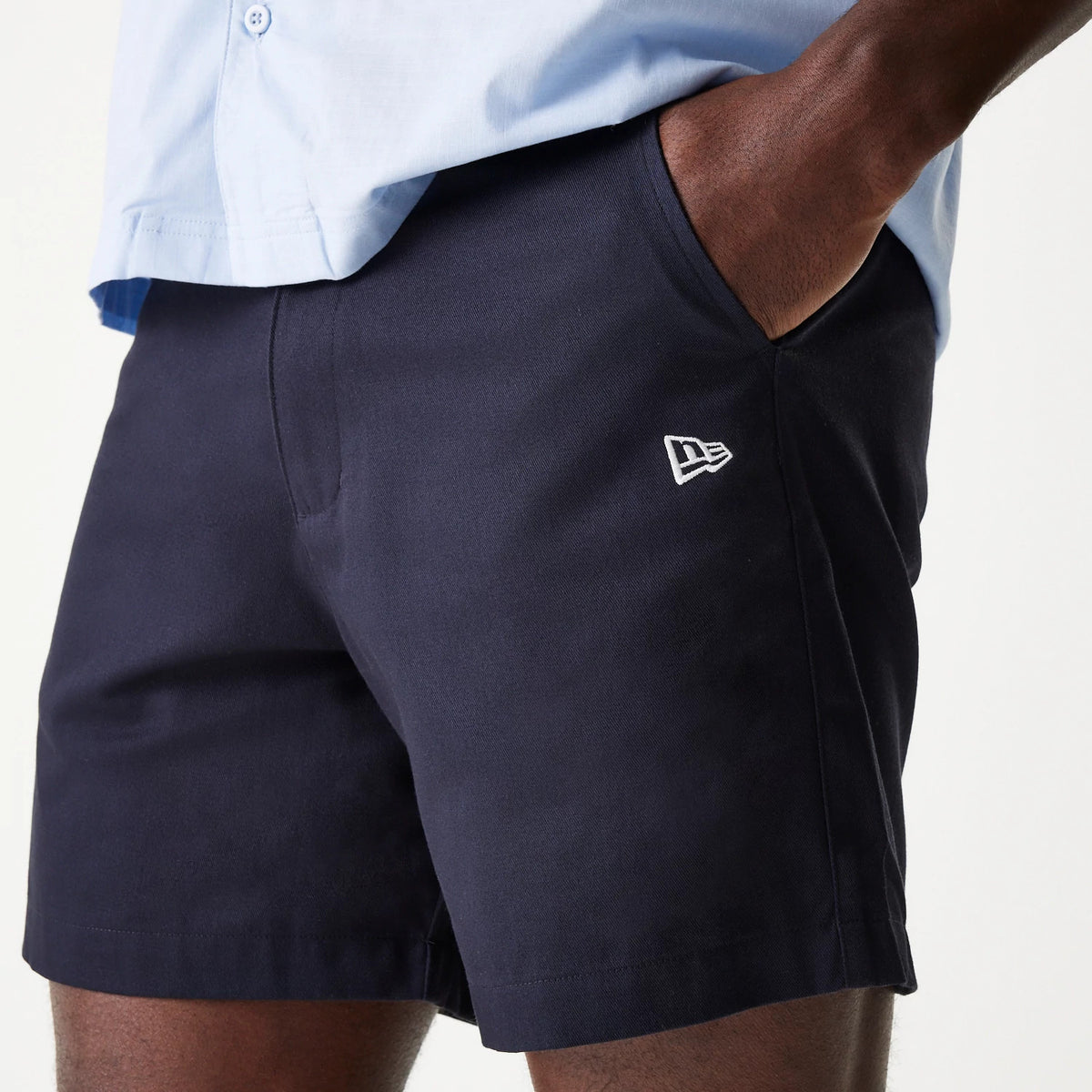 New Era Navy Chino Shorts, on-model view showing the navy shorts with a raised white New Era logo on the left leg, close-fitting around the thighs, and paired with a light blue shirt.