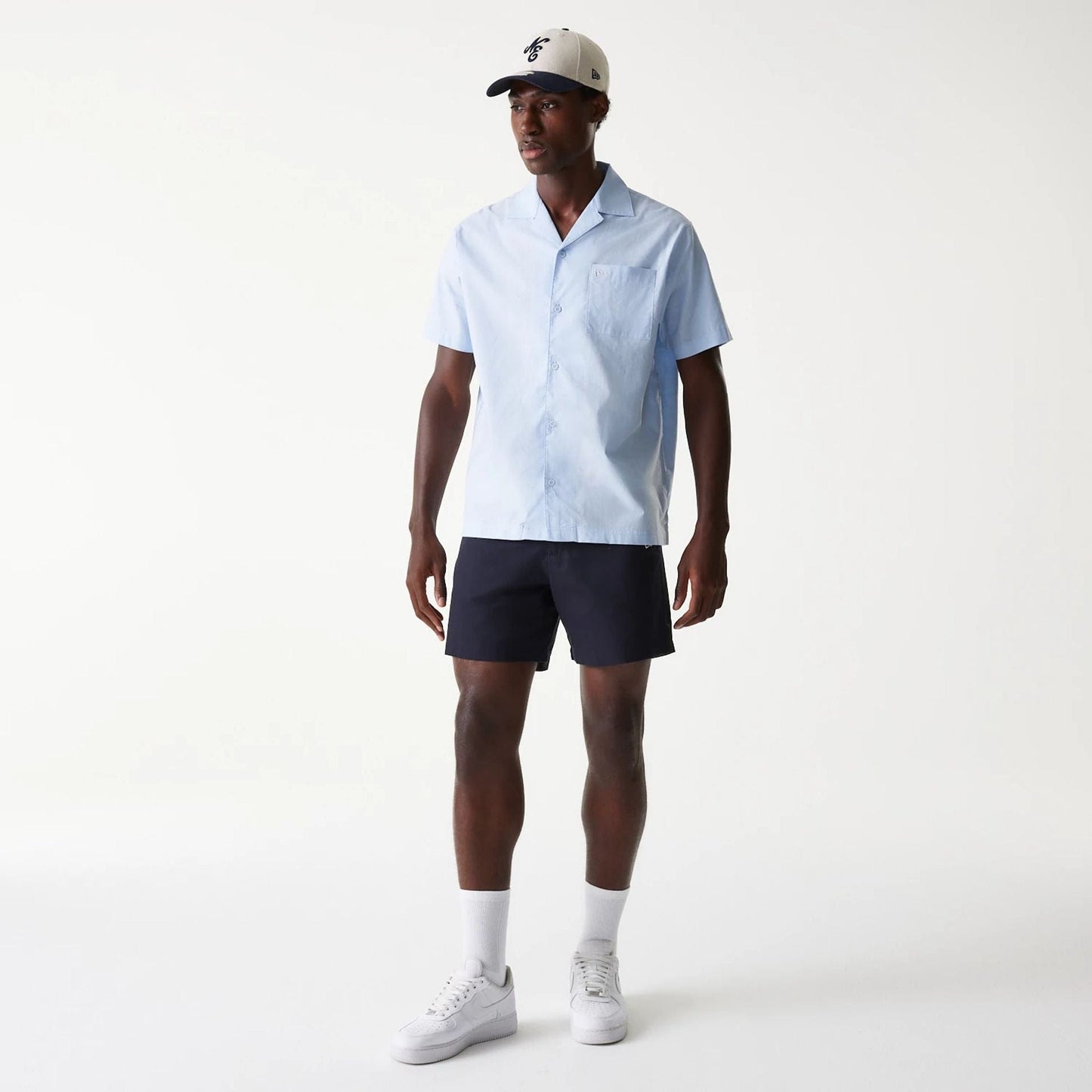 New Era Navy Chino Shorts, on-model view showing the dark navy shorts worn with a light blue shirt and white trainers, a debossed New Era logo on the front of a white cap with a black visor worn by the model.