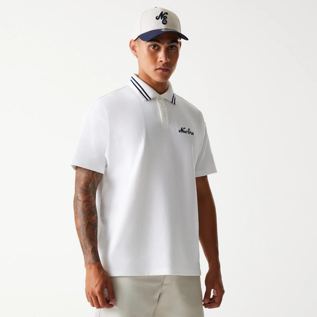 New Era Drop Shoulder White Polo Shirt, front view on a model with an embroidered New Era logo on the left chest, contrasting striped collar, short sleeves, and a model wearing a cap with an embroidered front logo.