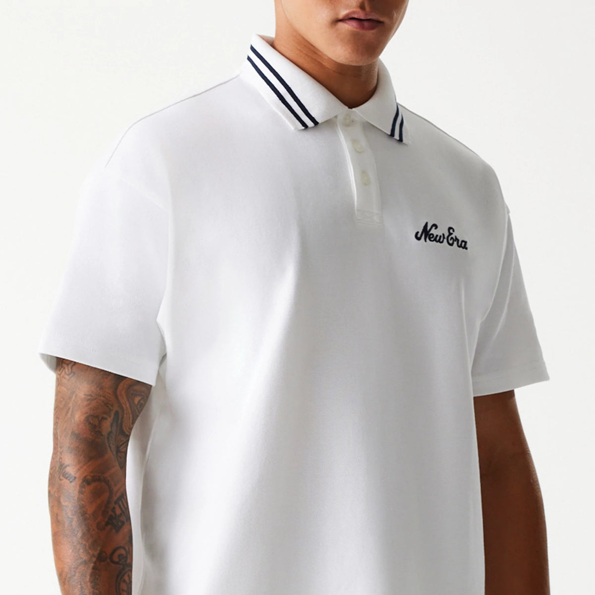 New Era Drop Shoulder White Polo Shirt, front view on a model showcasing the embroidered New Era logo in black on the left chest and a ribbed collar with navy stripes, paired with a drop shoulder design for a relaxed fit.