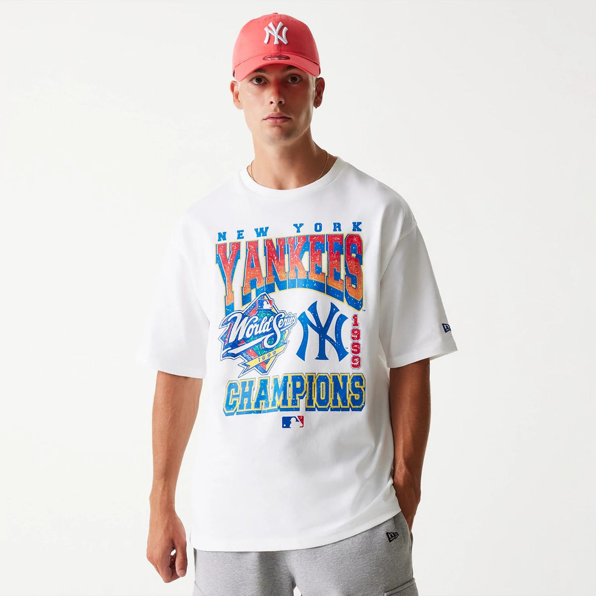 New Era New York Yankees MLB World Series 1999 White Oversized T-Shirt, front view featuring large colourful graphics including the New York Yankees logo and World Series emblem, worn by a model also wearing a pink cap with a raised NY logo.