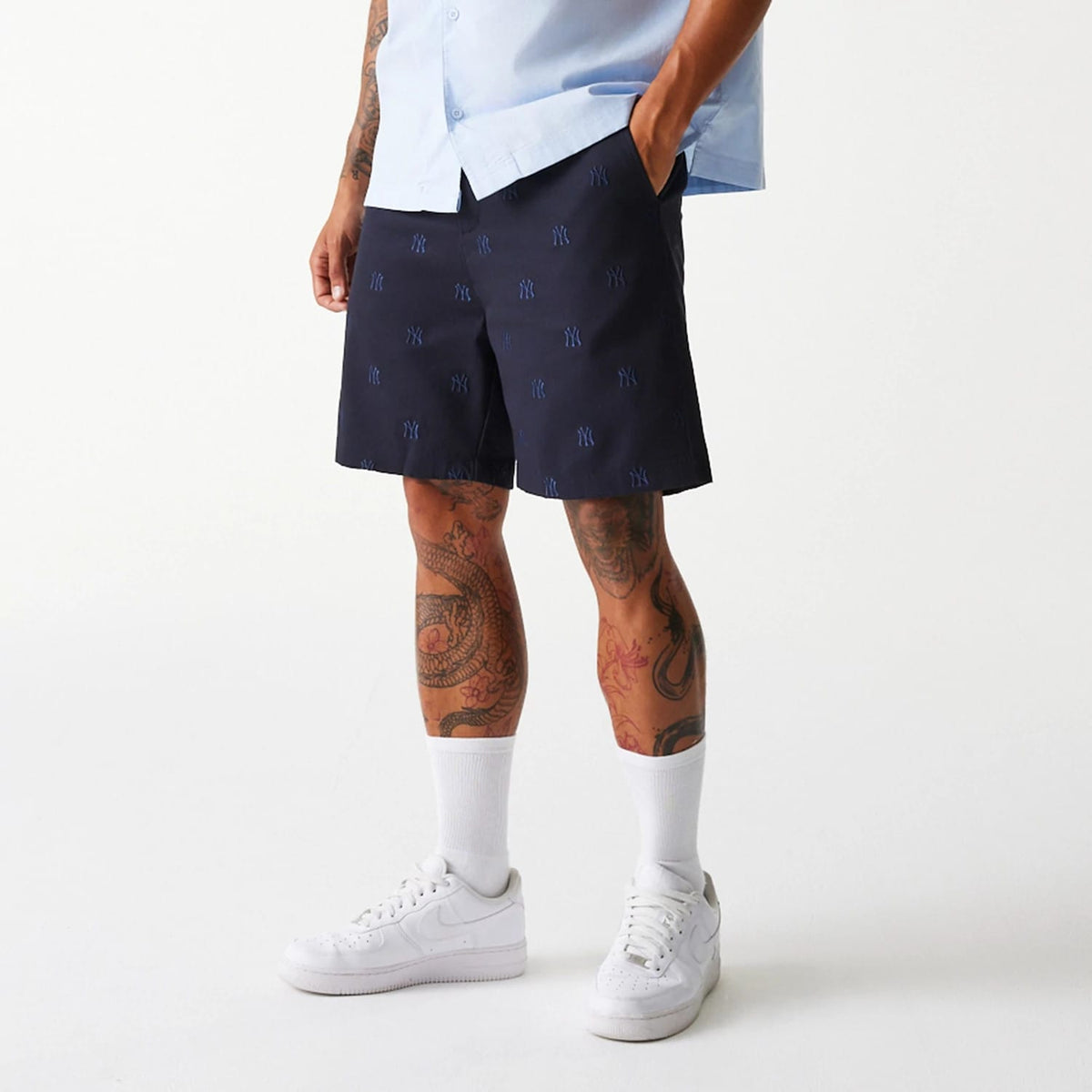 New Era New York Yankees MLB Monogram Navy Shorts, front view showing the navy fabric adorned with a pattern of raised New York Yankees logos throughout, worn by a model standing with hands in pockets, paired with white trainers and socks.