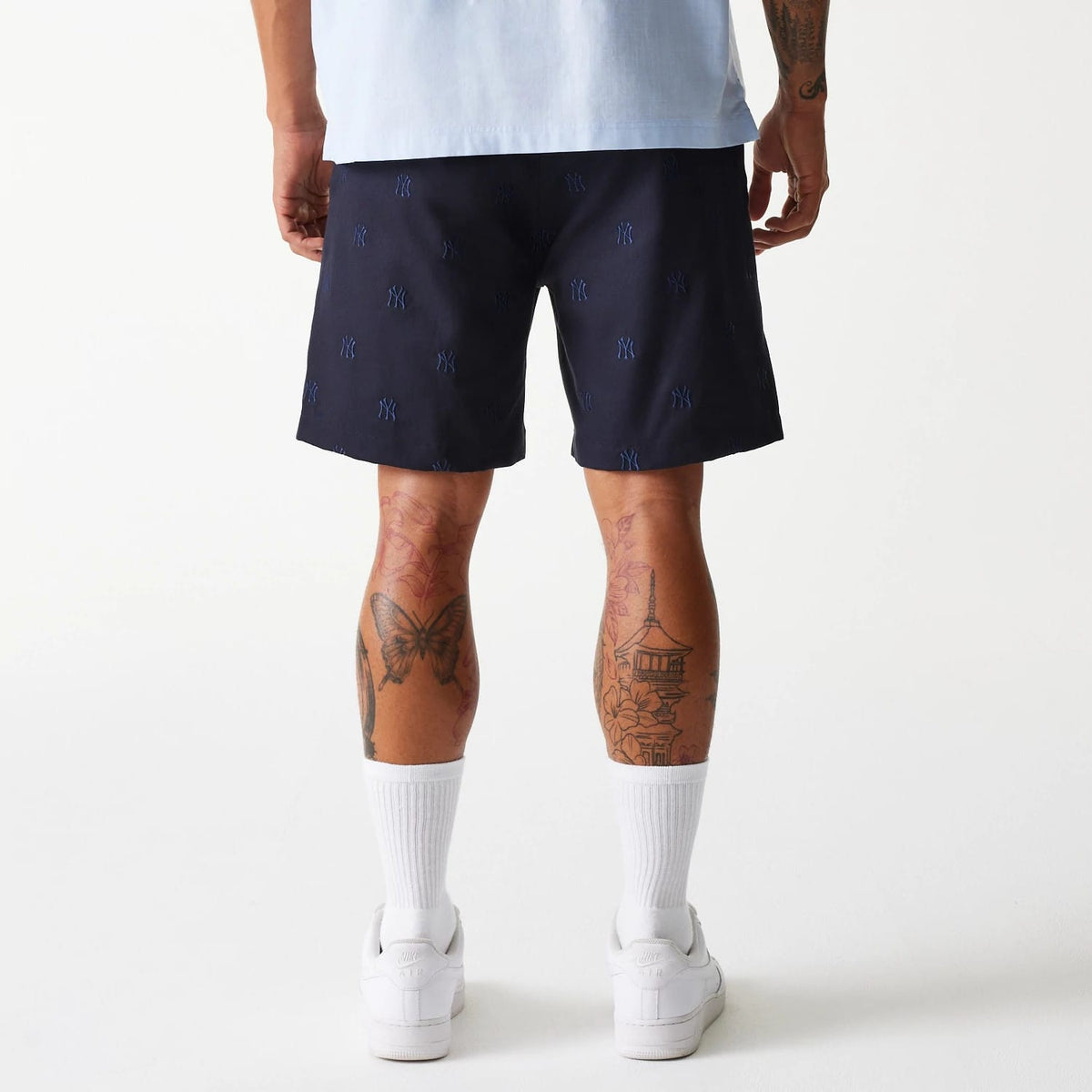 New Era New York Yankees MLB Monogram Navy Shorts, rear view showing navy blue shorts with an all-over raised New York Yankees logo pattern, worn by a tattooed individual standing with white sneakers and socks.