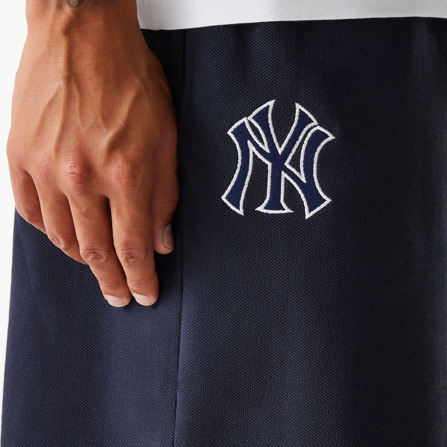 New Era New York Yankees MLB Pique Navy Shorts, side view showing the embroidered Yankees logo in white on the dark navy fabric, with part of a person's hand resting near the logo.