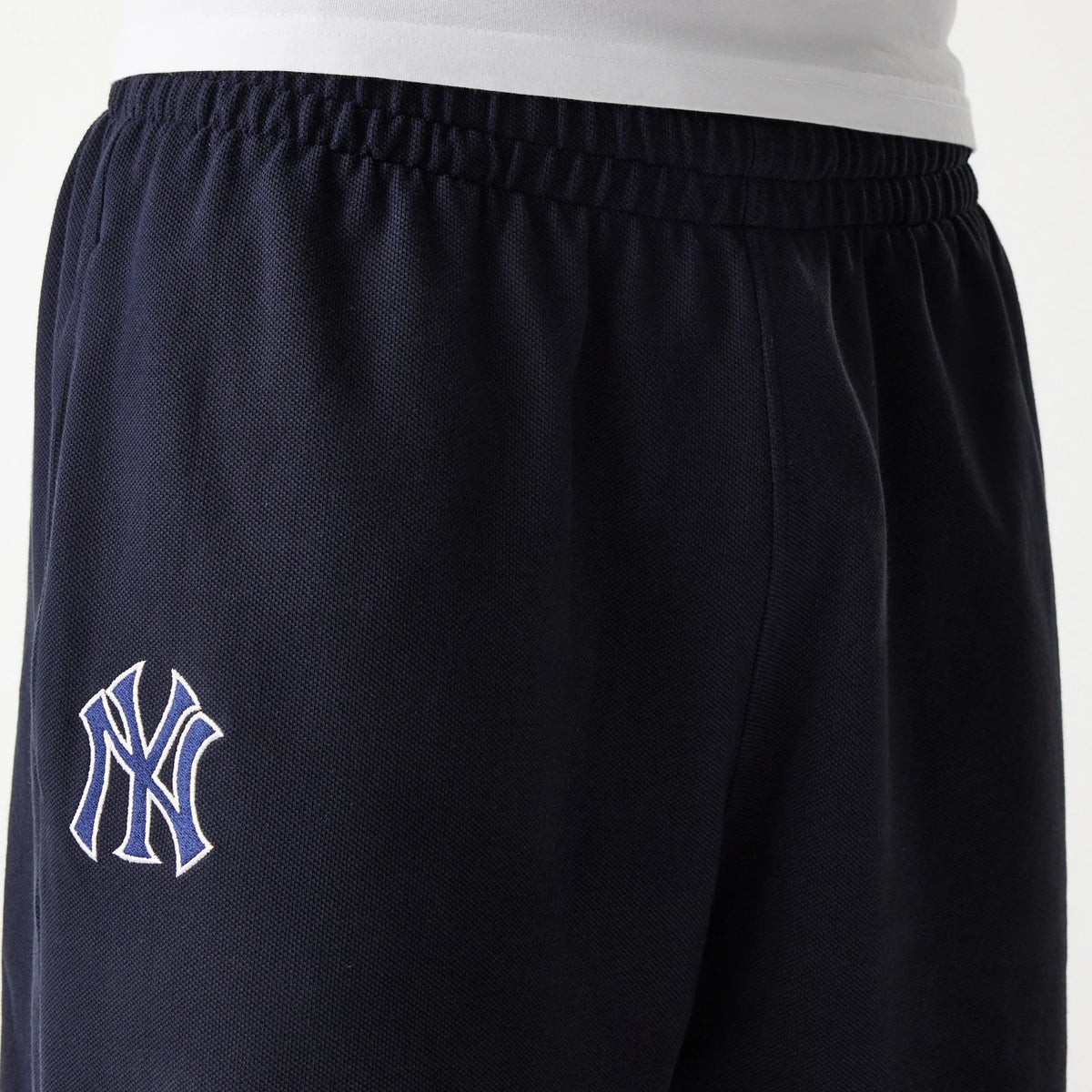 New Era New York Yankees MLB Pique Navy Shorts, side view showing the elastic waistband at the top, the navy fabric with a raised embroidered New York Yankees logo in white and blue on the left leg, and a glimpse of a white shirt above the shorts.