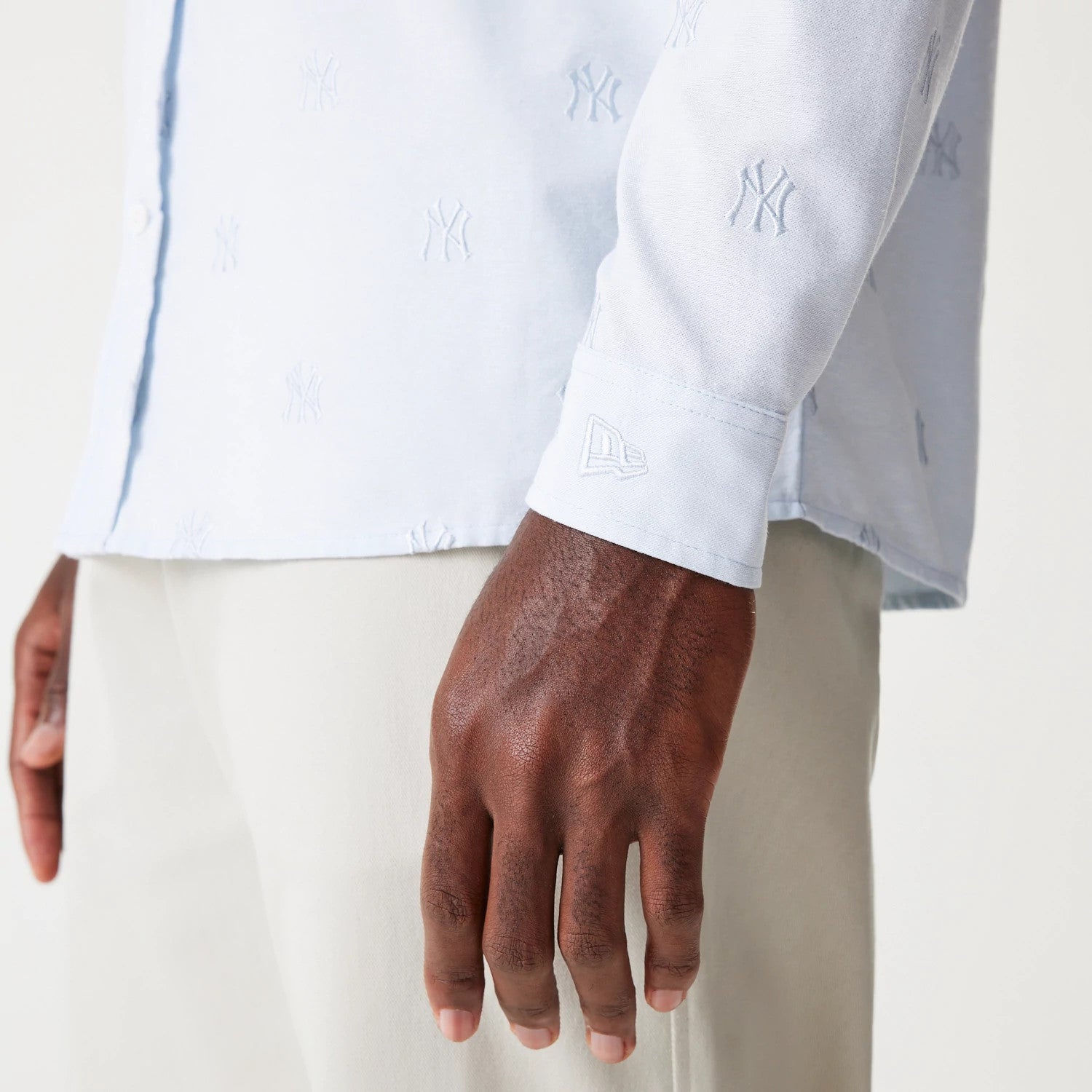 New Era New York Yankees MLB Monogram Pastel Blue Long Sleeve Shirt, side view on a model showing white embroidered NY logos across the fabric, with a New Era flag logo on the cuff and light trousers complementing the design.