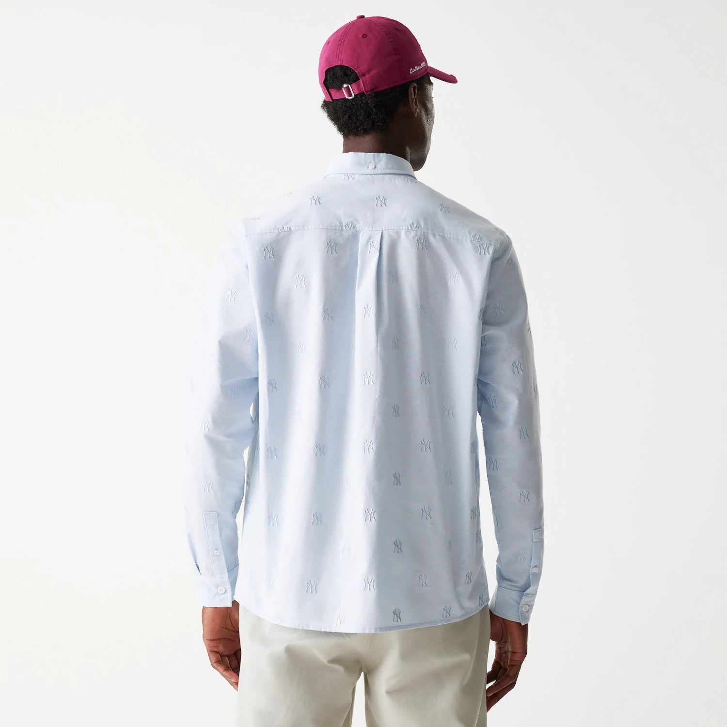 New Era New York Yankees MLB Monogram Pastel Blue Long Sleeve Shirt, rear view on a model wearing a red cap, showing the embroidered NY monogram pattern across the light blue fabric and buttoned cuffs.