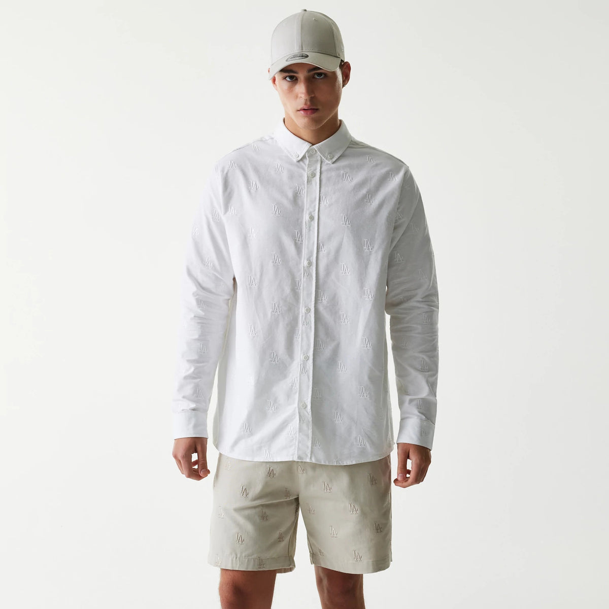 New Era LA Dodgers MLB Monogram White Long Sleeve Shirt, front view on a model wearing matching beige shorts and a light grey cap, showing repeated LA logo embroidery across the shirt's front and sleeves.
