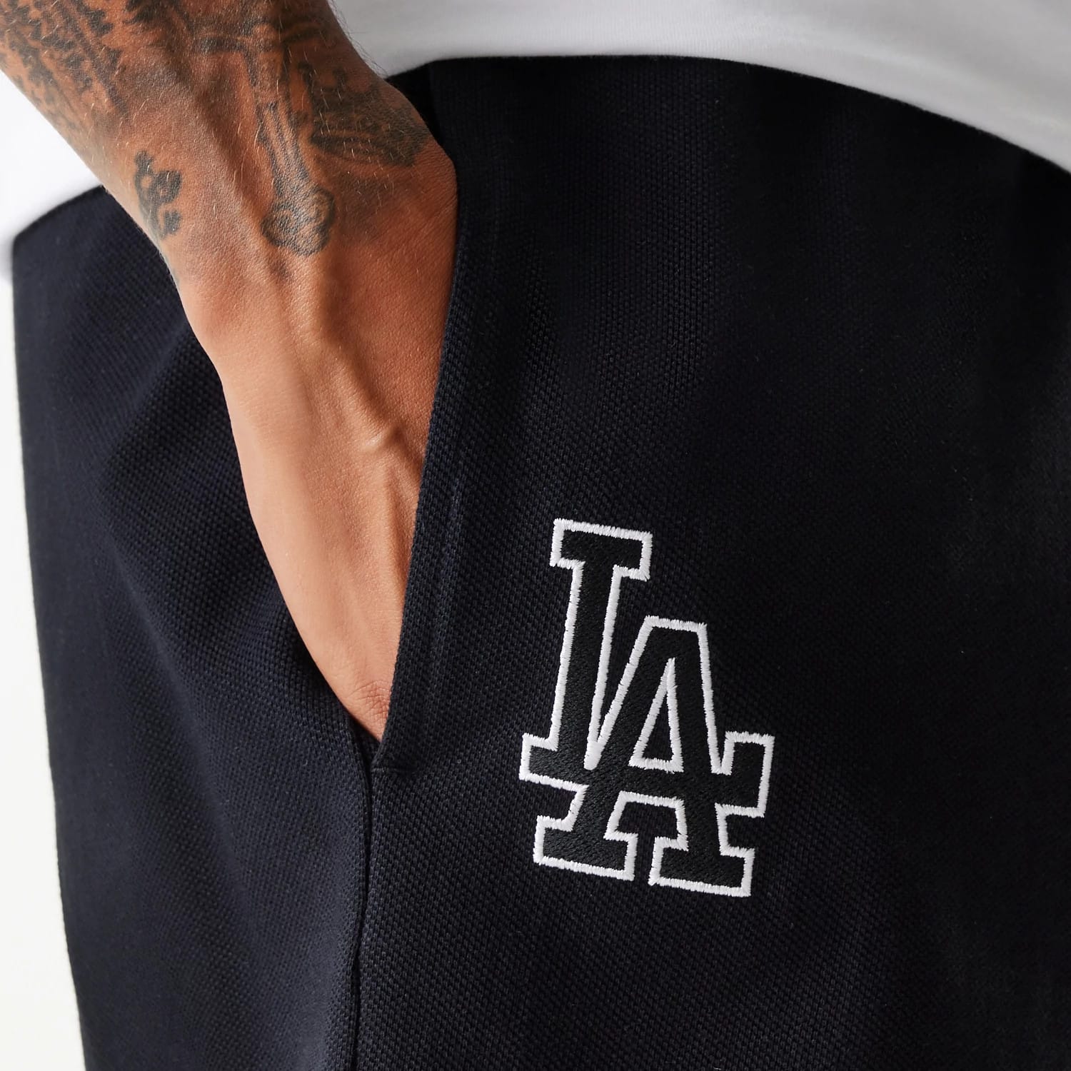 New Era LA Dodgers MLB Pique Black Shorts, front view showing a large white embroidered LA logo on the right leg near the pocket, with a person's hand partially tucked into the pocket against the black fabric.