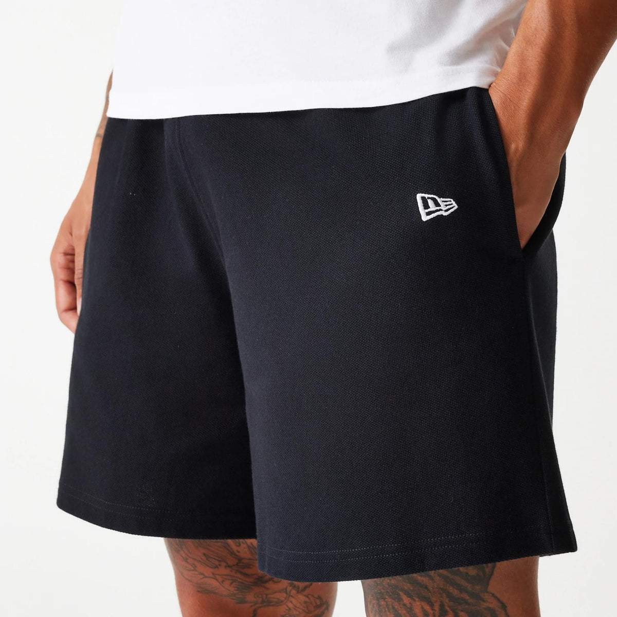 New Era LA Dodgers MLB Pique Black Shorts, on-model view showing the black shorts with an embroidered white New Era logo on the left leg, contrasting against the dark fabric, with one hand in the side pocket.