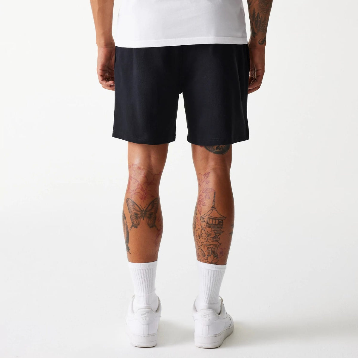New Era LA Dodgers MLB Pique Black Shorts, rear view showing the simple black design reaching above the knees, with a relaxed fit, no visible logos or patterns, worn by a model with tattooed legs and white trainers.