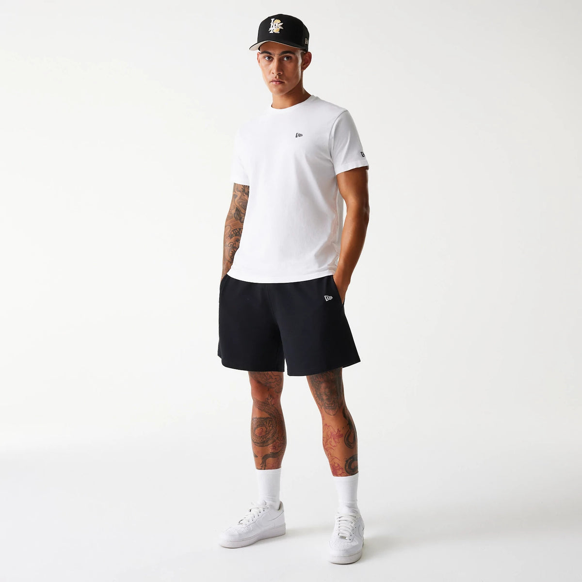 New Era LA Dodgers MLB Pique Black Shorts, on-model view showing a black New Era logo on the left leg of the black shorts, paired with a white t-shirt featuring a small black debossed New Era logo on the chest and a black cap.