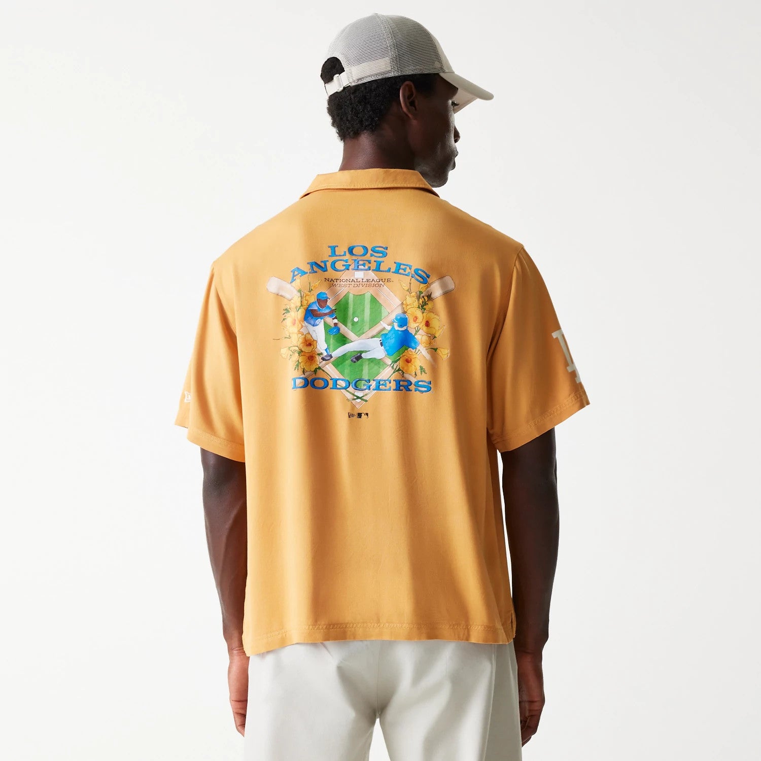 New Era LA Dodgers MLB Graphic Beige Revere Short Sleeve Shirt, rear view on a model wearing white trousers and a light cap, featuring a colourful graphic of two players, flowers, and baseball bats with blue Los Angeles Dodgers lettering.