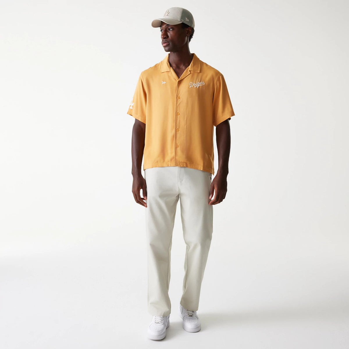 New Era LA Dodgers MLB Graphic Beige Revere Short Sleeve Shirt, on a model styled with white trousers and trainers, featuring white embroidered Dodgers script on the left chest and sleeve, and clean front button closure.