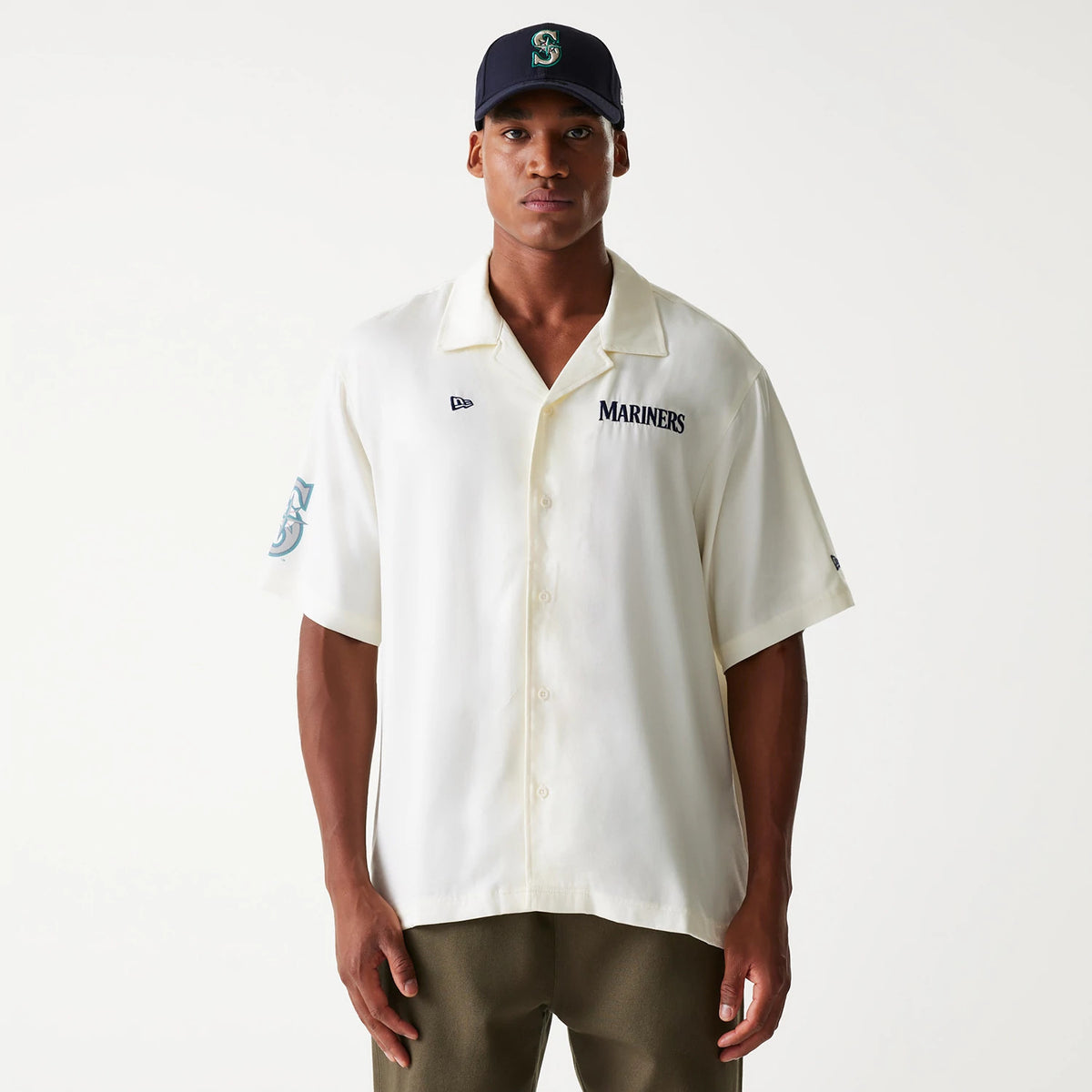 New Era Seattle Mariners MLB Graphic Open White Revere Short Sleeve Shirt, front view on a model wearing a black cap with the Mariners S logo, featuring embroidered Mariners lettering on the right chest and a teal Mariners logo on the left sleeve.