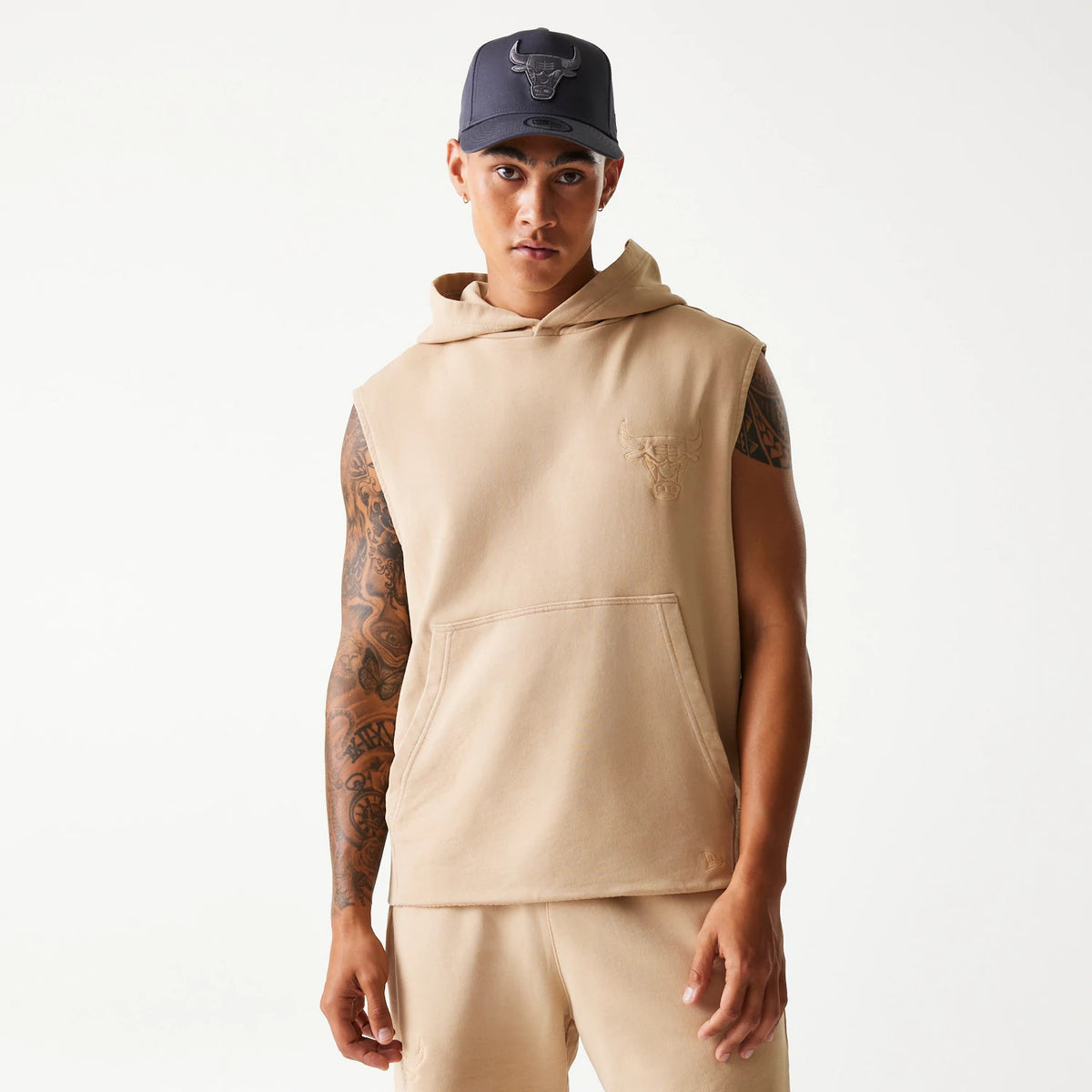 New Era Chicago Bulls NBA Washed Beige Sleeveless Hoodie, on-model view showing sleeveless design with a kangaroo pocket on the front, the raised Bulls logo on the left chest, and a hood at the back pairing with beige shorts.