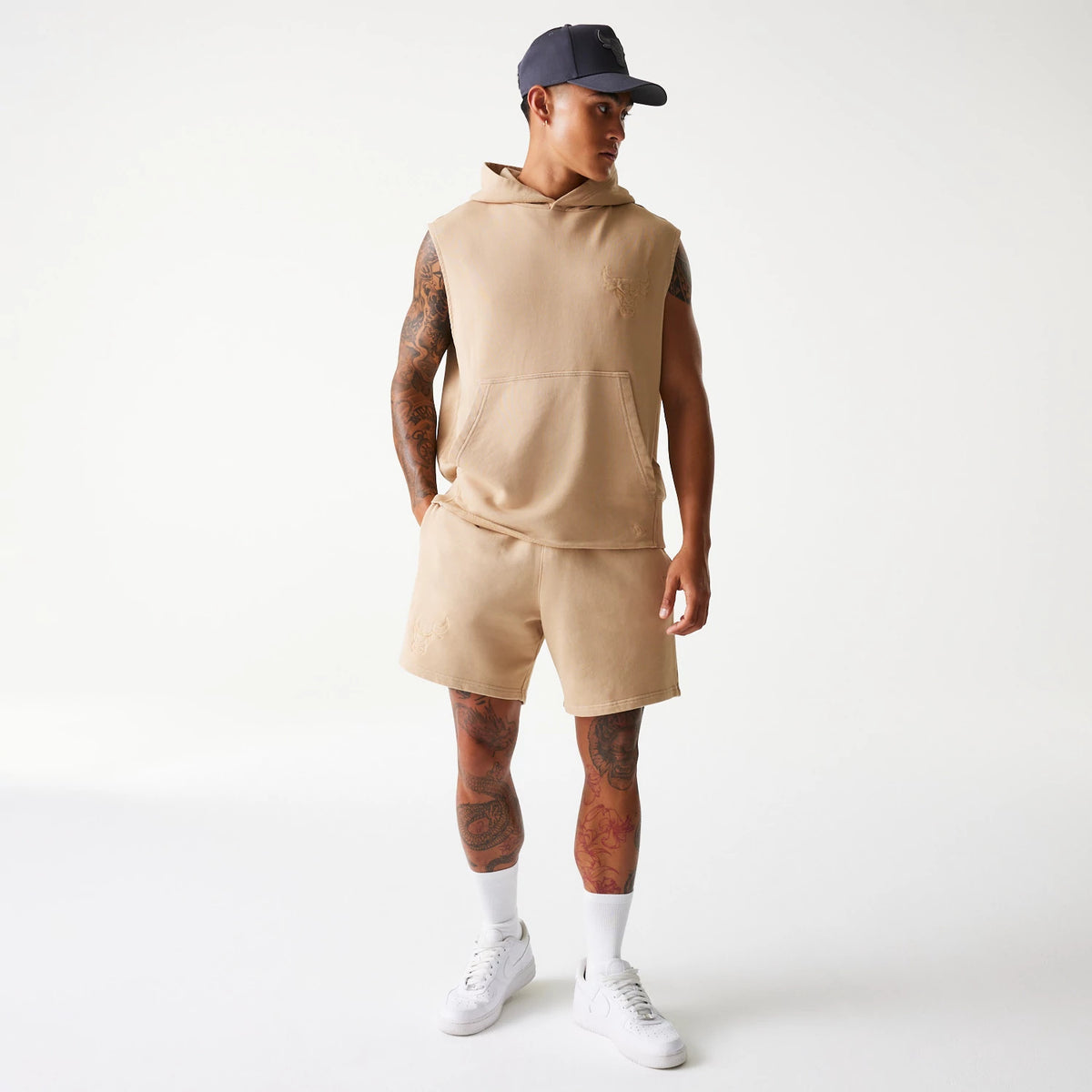 New Era Chicago Bulls NBA Washed Beige Sleeveless Hoodie, on-model view showing a front kangaroo pocket, an embossed Chicago Bulls logo on the left chest, and sleeveless cut paired with matching beige shorts and white trainers.