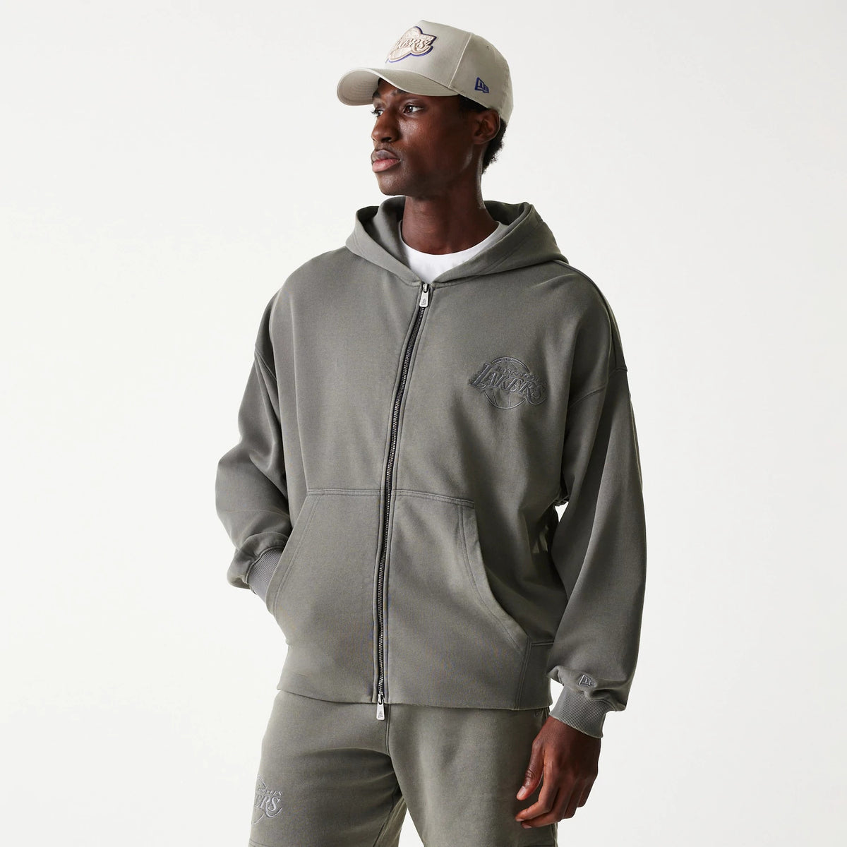 New Era LA Lakers NBA Washed Grey Full Zip Hoodie, on-model view showing the grey zip-up hoodie with a raised Lakers logo on the left chest, a hood, and ribbed cuffs, paired with matching grey trousers and a beige cap.