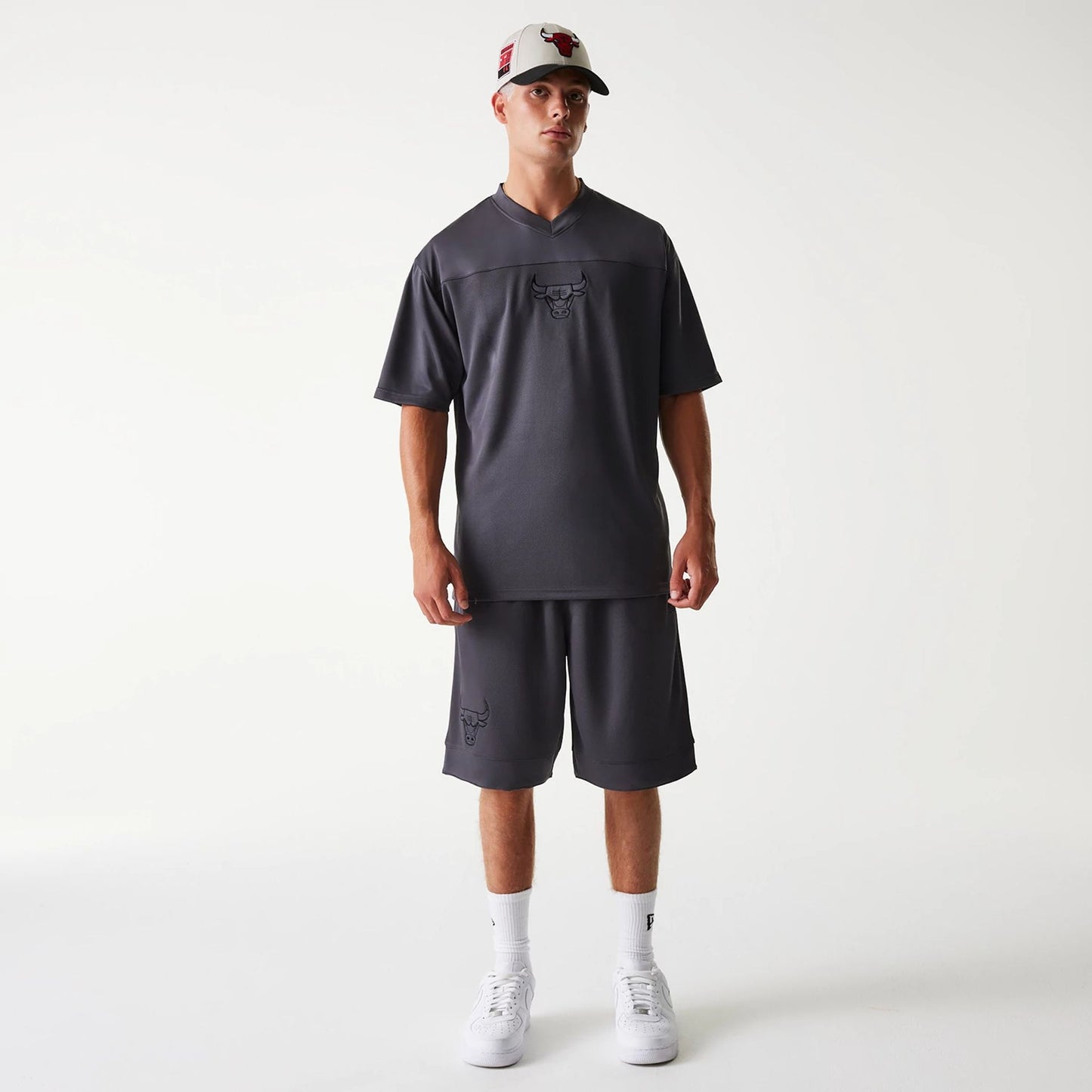 New Era Chicago Bulls NBA Tonal Dark Grey Mesh Jersey, on a model with matching dark grey shorts, centre front view showing the tonal Bulls logo on the chest and shorts, styled with white trainers and a white cap with a red Bull logo.