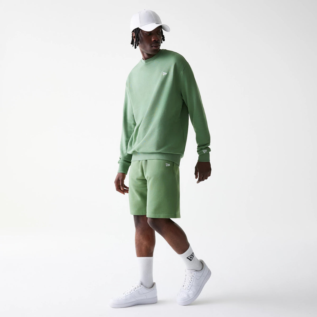 New Era Essential Green Oversized Shorts, on-model view showing the relaxed fit green shorts with a raised New Era logo on the left leg, paired with a matching green sweatshirt and white trainers.