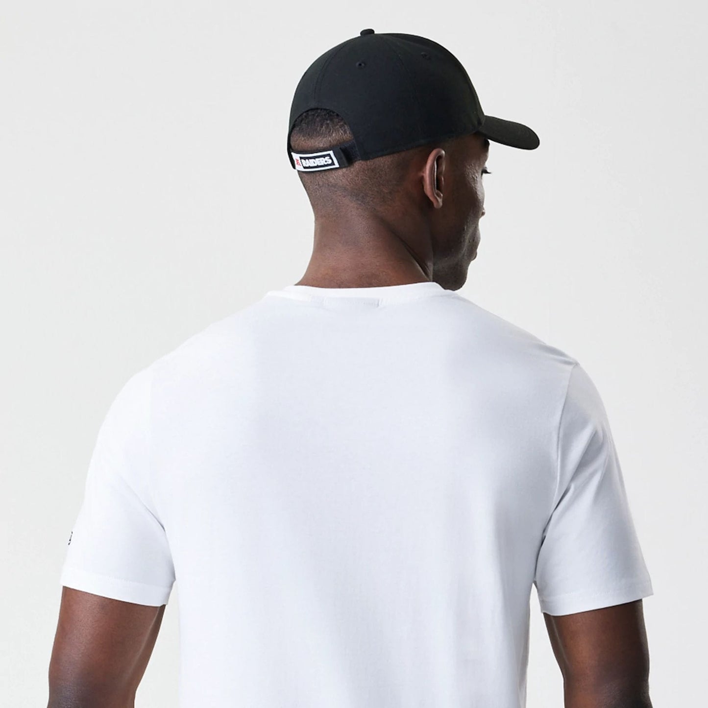 New Era Las Vegas Raiders NFL Stack Arch Logo White T-Shirt, rear view showing plain white back with short sleeves, on-model wearing a black cap with a logo on the strap and eyelets on top, head turned slightly to the side.