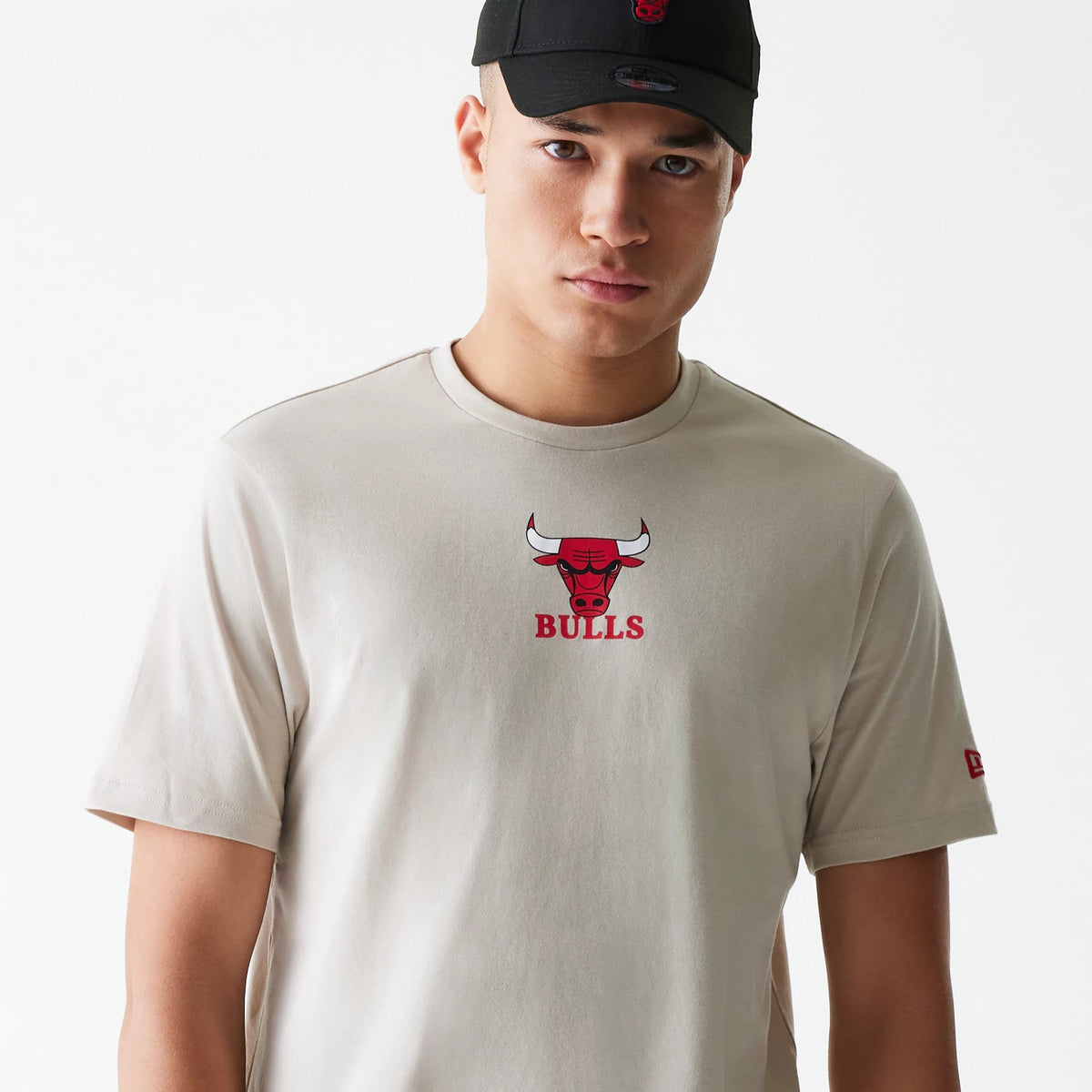New Era Chicago Bulls NBA Logo Cream T-Shirt, on-model view showing a cream T-shirt with a red Chicago Bulls logo centred on the chest, the New Era logo in red debossed on the left sleeve, and a black cap on the model.