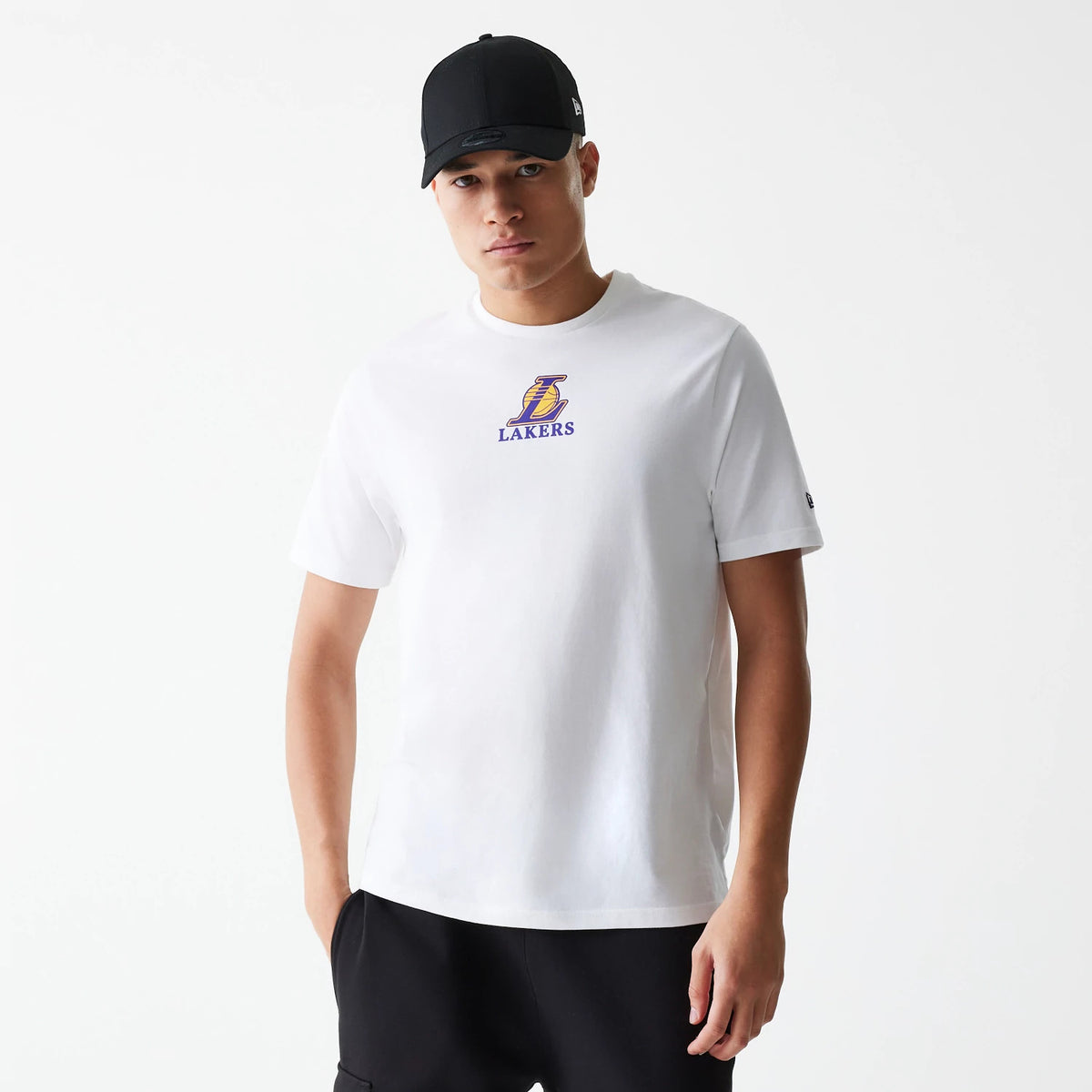 New Era LA Lakers NBA Logo White T-Shirt, on-model view showing the white T-shirt with the Lakers logo in purple and yellow centred on the chest, worn by a person in black trousers and a black cap with a debossed New Era logo on the side.