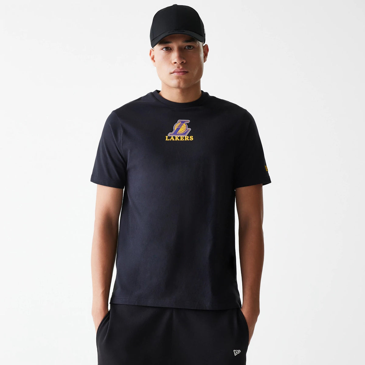 New Era LA Lakers NBA Logo Black T-Shirt, front view showing a raised Lakers logo in purple and gold centred on the chest, worn by a model with a plain black cap, short sleeves, and a branded badge on the left sleeve.