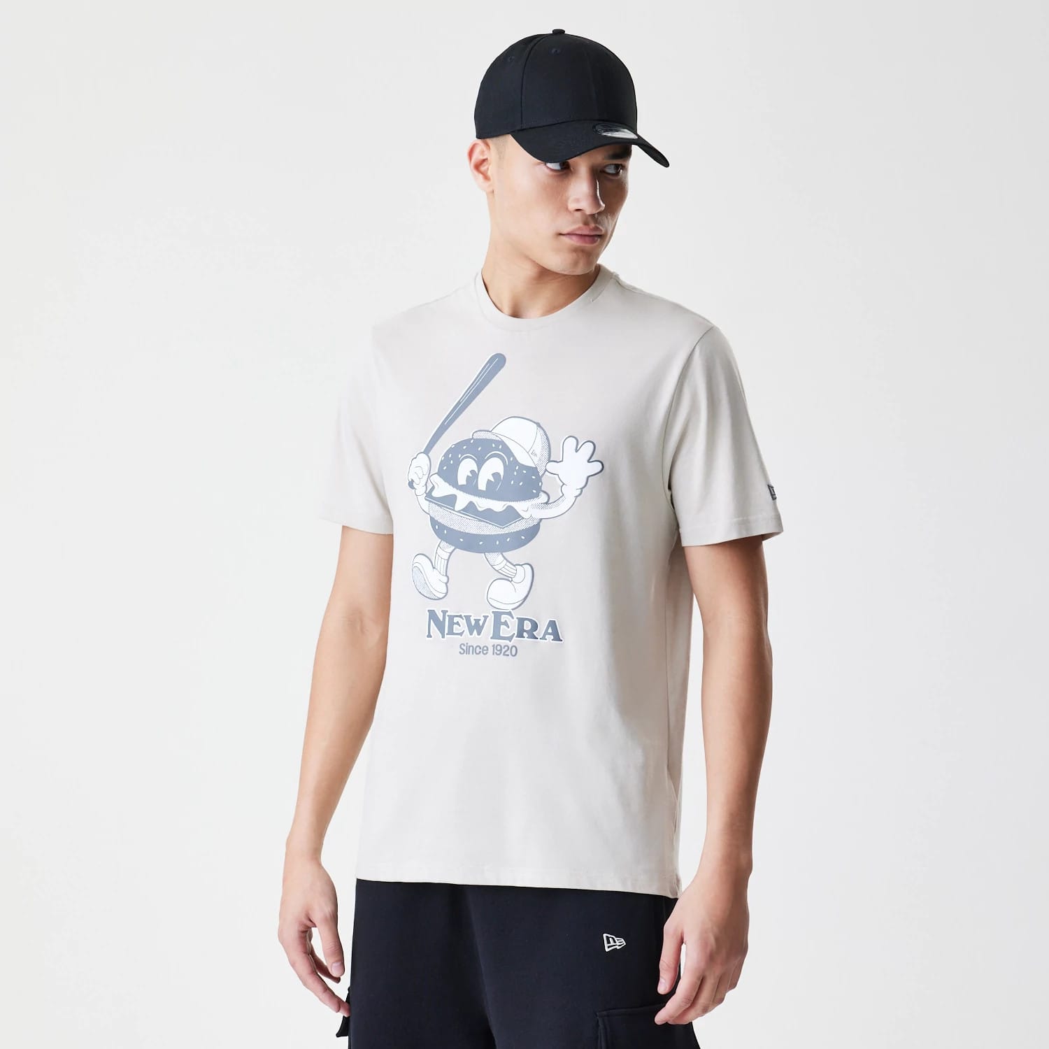 New Era Burger Graphic Cream T-Shirt, on-model view showing the cream shirt with a large cartoon burger graphic on the front, featuring a debossed New Era logo beneath and a man wearing a black cap.