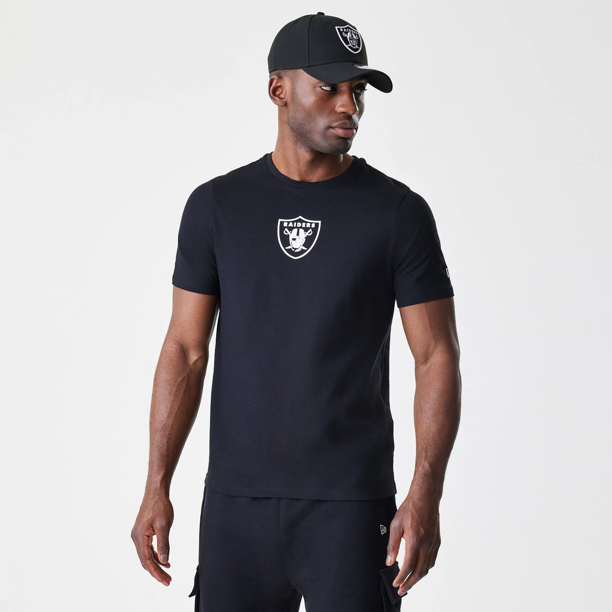 New Era Las Vegas Raiders NFL Logo Black T-Shirt, on-model view showing the short-sleeved black T-shirt with a raised white Raiders logo centred on the chest, paired with a black cap featuring the same Raiders logo on the front.