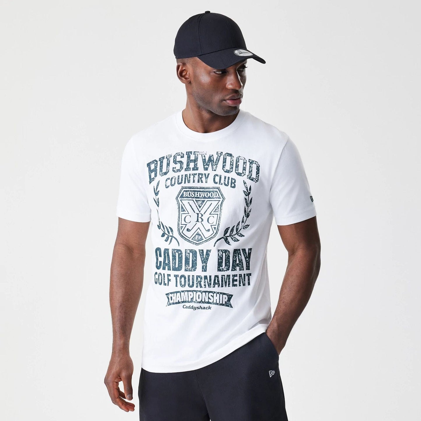 New Era Caddyshack Caddy Day Tournament White T-Shirt, front view showing bold black text and a crest on the chest, worn by a person also wearing a dark cap with a debossed New Era logo on the side of the cap.