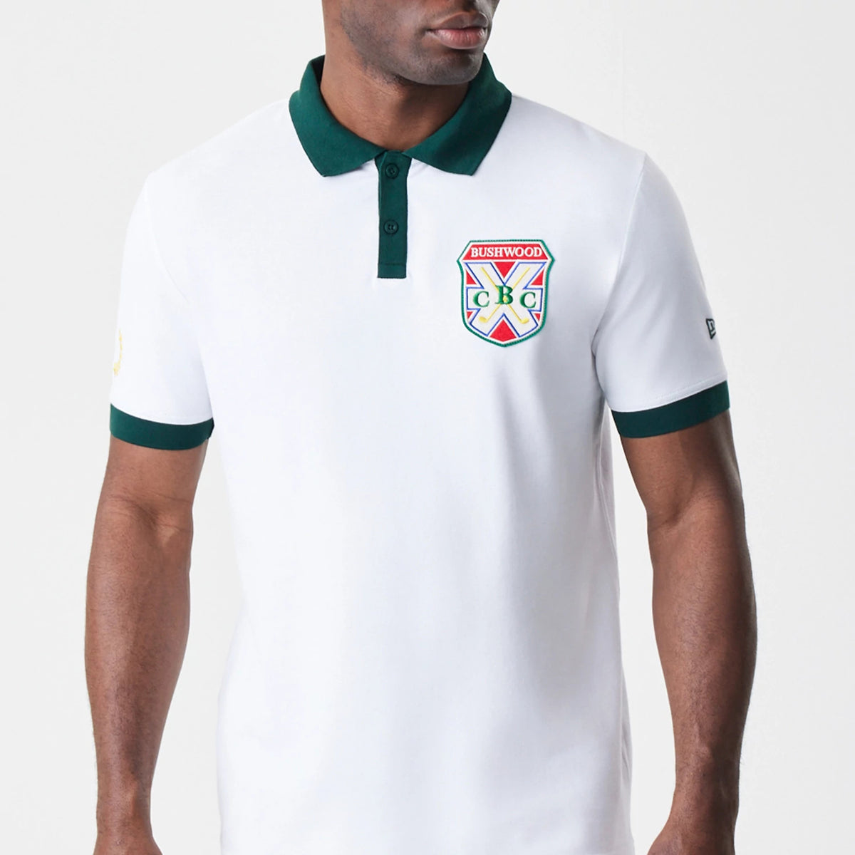 New Era Caddyshack BushWood White Polo T-Shirt, front view showing the white shirt with a green collar and sleeve edges, an embroidered BushWood crest on the left chest, and a small debossed New Era logo on the left sleeve.