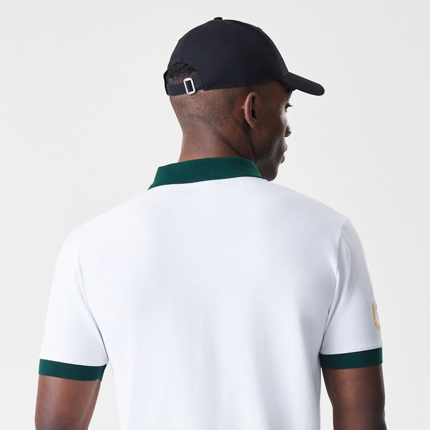 New Era Caddyshack BushWood White Polo T-Shirt, rear view showing a plain white back with dark green cuffs and collar, worn by a model in a black cap with a silver buckle on the strap, highlighting simple elegance and contrasting details.