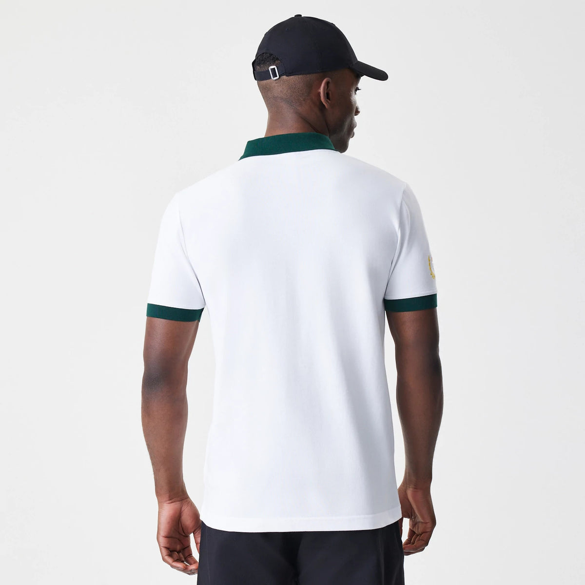 New Era Caddyshack BushWood White Polo T-Shirt, rear view showing a plain white back with dark green collar and sleeve cuffs, a small raised logo on the left sleeve, and the shirt worn by a model with a black cap.