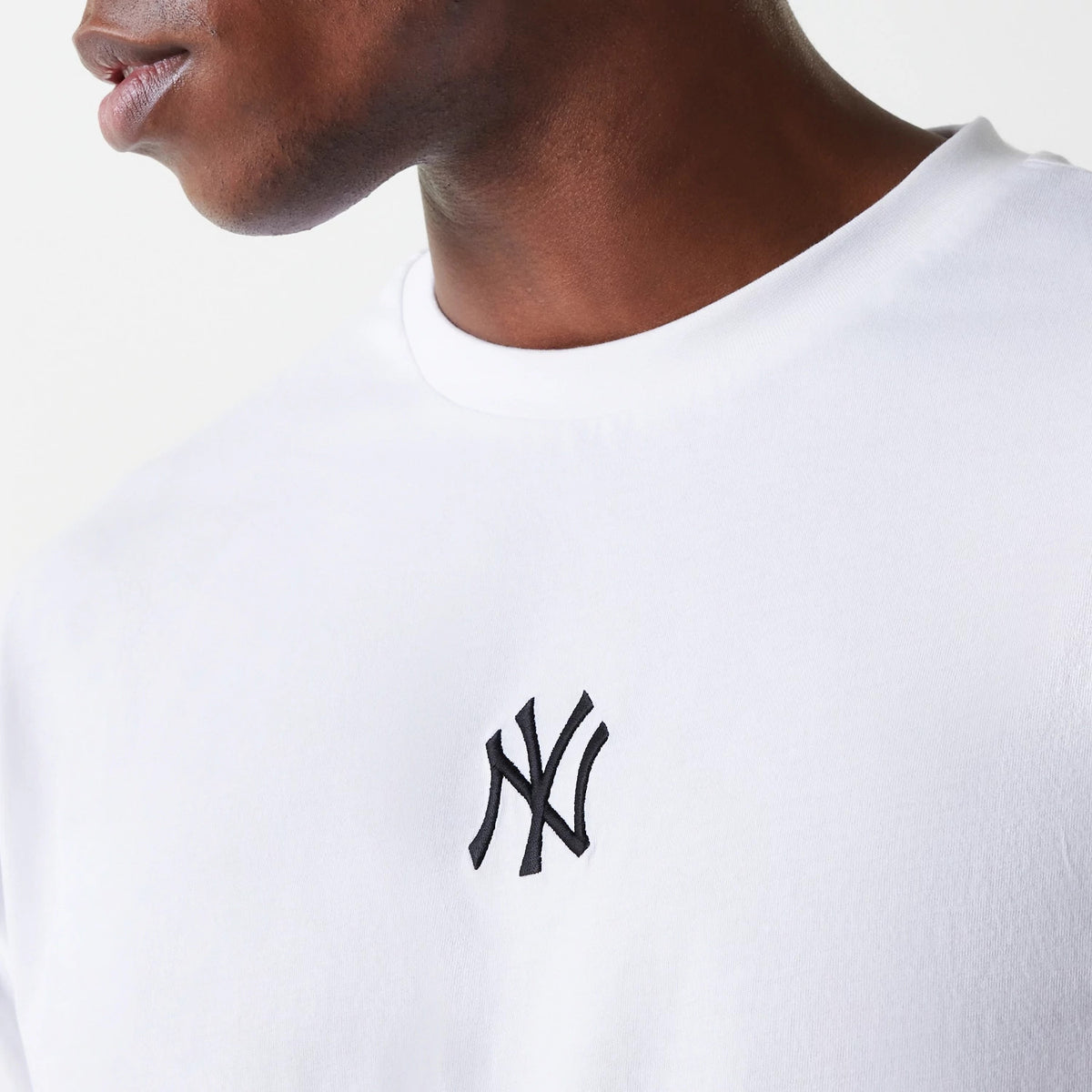 New Era New York Yankees MLB League Essential White Oversized T-Shirt, on-model view showing a front close-up of the white fabric with a black embroidered New Era logo on the chest, worn by a person turned slightly to the side.