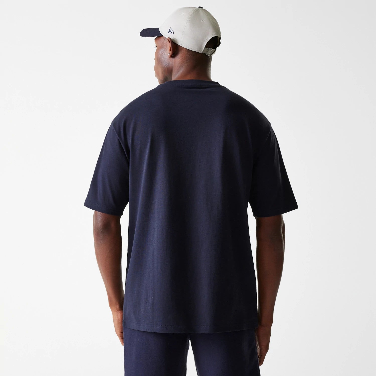 New Era New York Yankees MLB League Essential Navy Oversized T-Shirt, rear view showing the plain navy back of the oversized T-shirt worn by a model, paired with a light-coloured cap having the New Era debossed logo on the side.