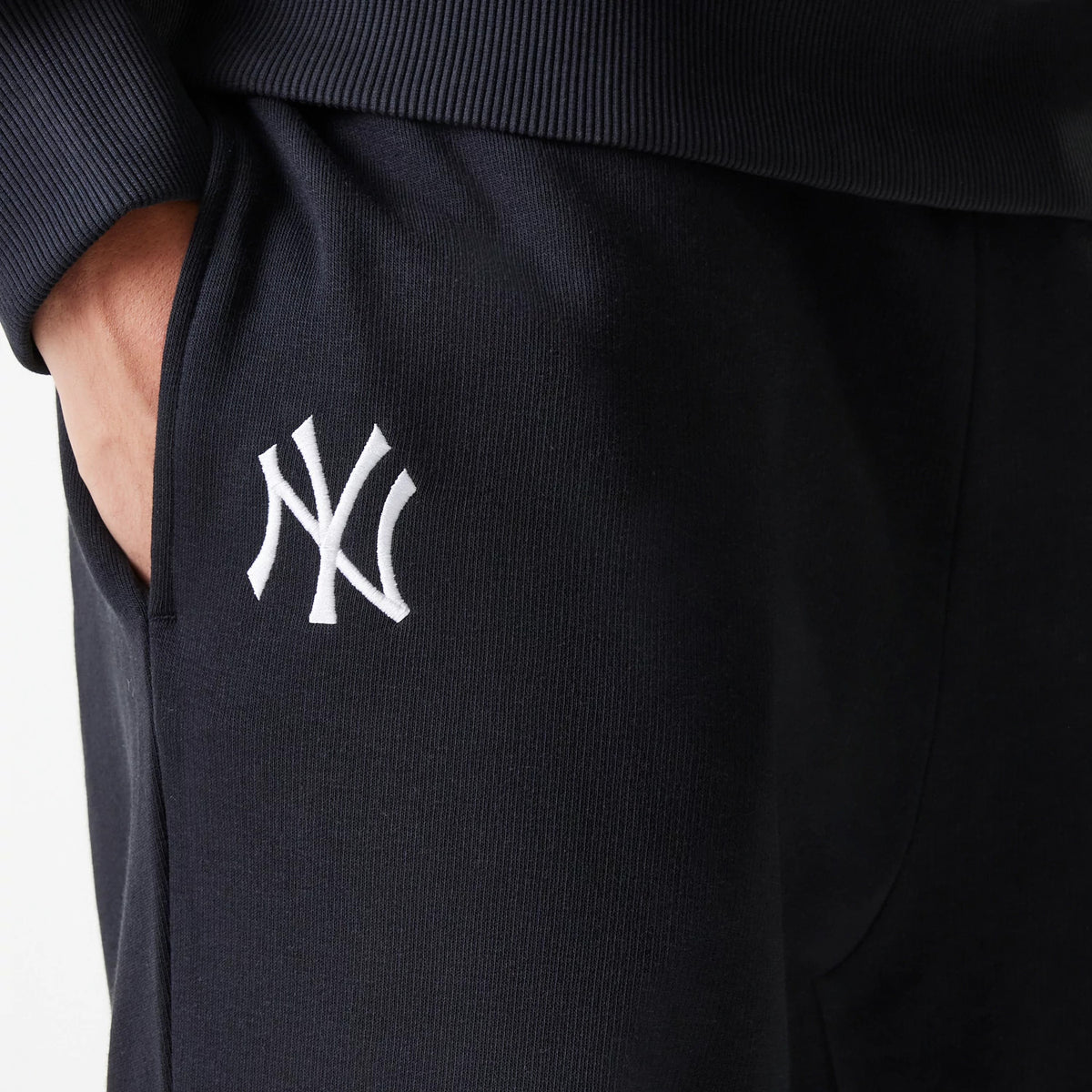 New Era New York Yankees MLB League Essential Midi French Terry Black Joggers, close-up showing the white embroidered NY logo on the left thigh, with a hand in the pocket and ribbed waistband detail.