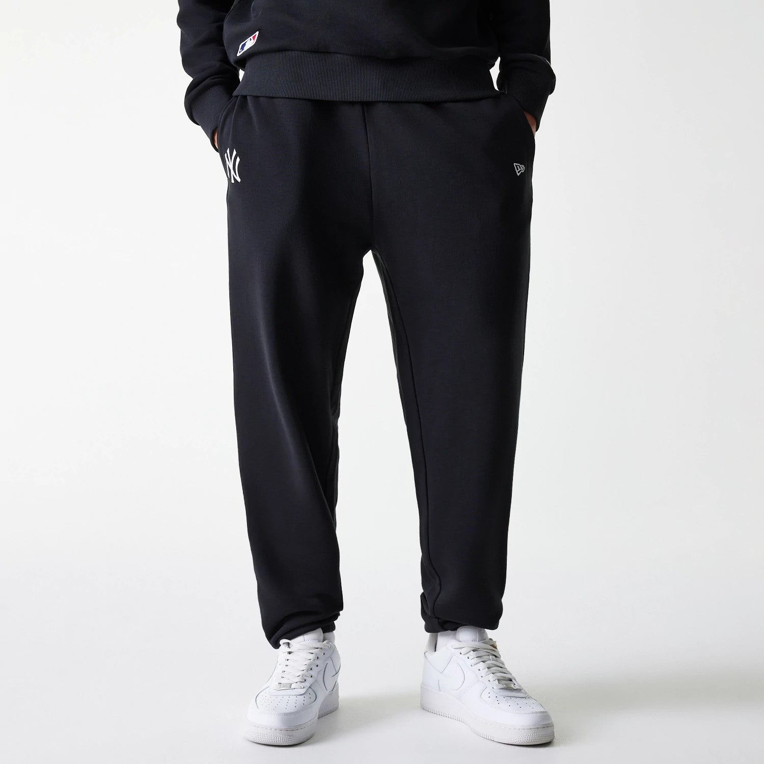New Era New York Yankees MLB League Essential Midi French Terry Black Joggers, front view on a model showing the white embroidered NY logo on the left leg, black New Era flag logo on the pocket, and paired with white trainers.