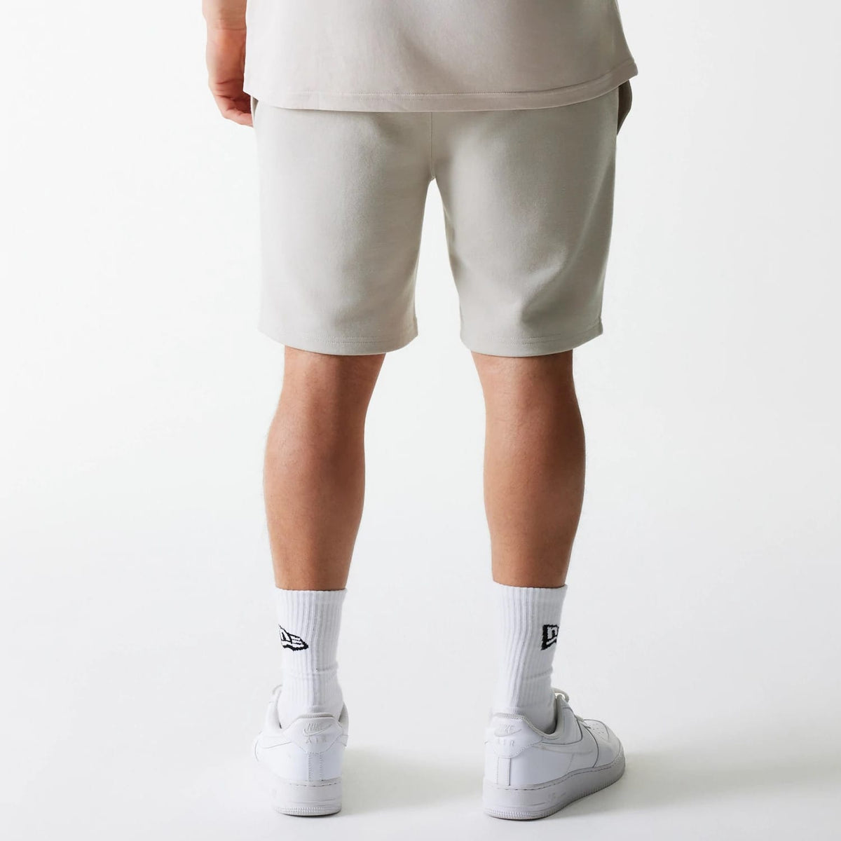 New Era New York Yankees MLB League Essential French Terry Cream Shorts, rear view showing the cream colour shorts worn by a model, with the hem resting just above the knee, and white socks with a black embroidered New Era logo above white shoes.