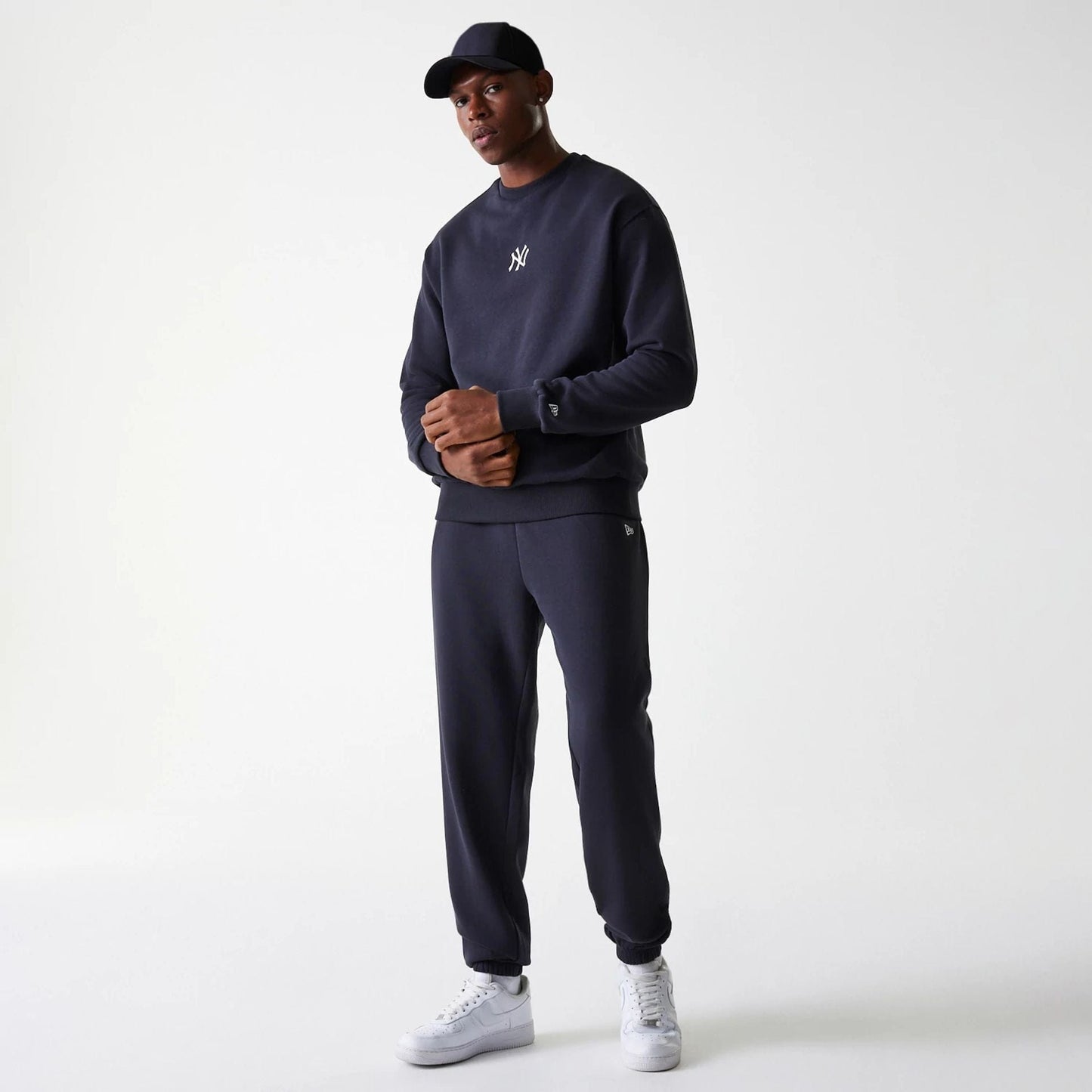 New Era New York Yankees MLB League Essential French Terry Navy Crew Neck Sweater, on a model styled with matching navy joggers and a black cap, featuring the white Yankees logo on the chest and the New Era flag logo on the left sleeve.