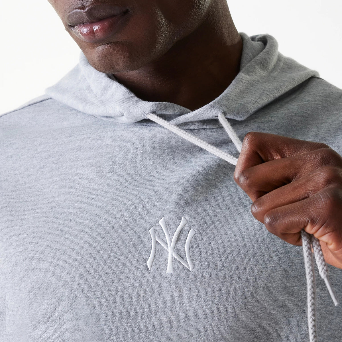 New Era New York Yankees MLB League Essential Midi French Terry Grey Oversized Pullover Hoodie, front view showing the embossed white NY Yankees logo on the chest, with a drawstring hood pulled slightly by hand, highlighting the soft grey fabric.