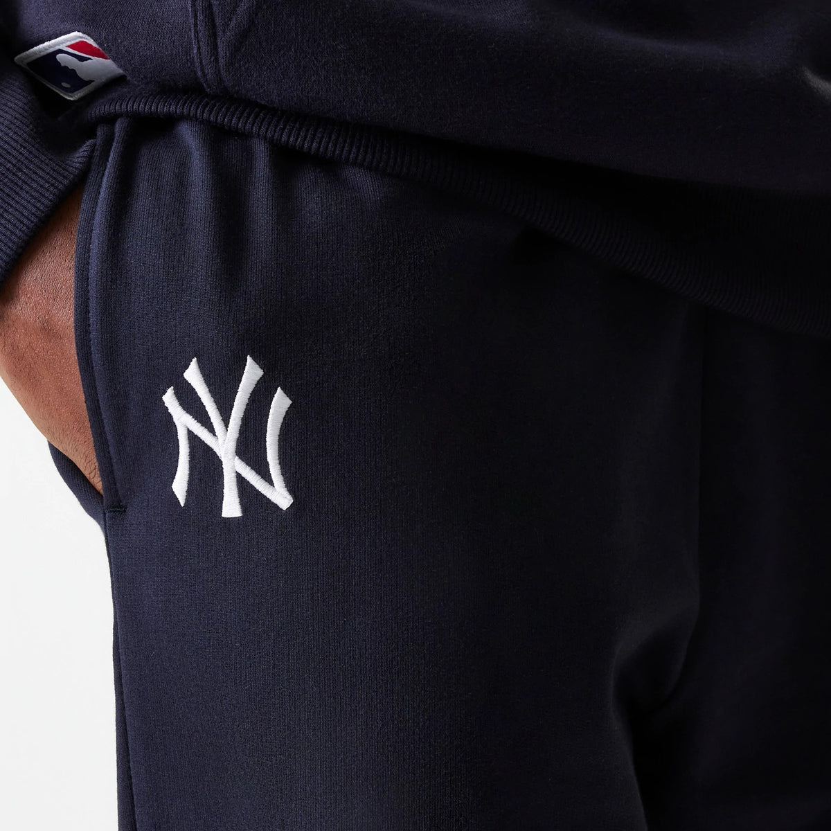 New Era New York Yankees MLB League Essential Midi French Terry Navy Joggers, close-up showing the white embroidered NY logo on the left thigh and the MLB Batterman logo on the ribbed hem, highlighting the soft terry fabric.
