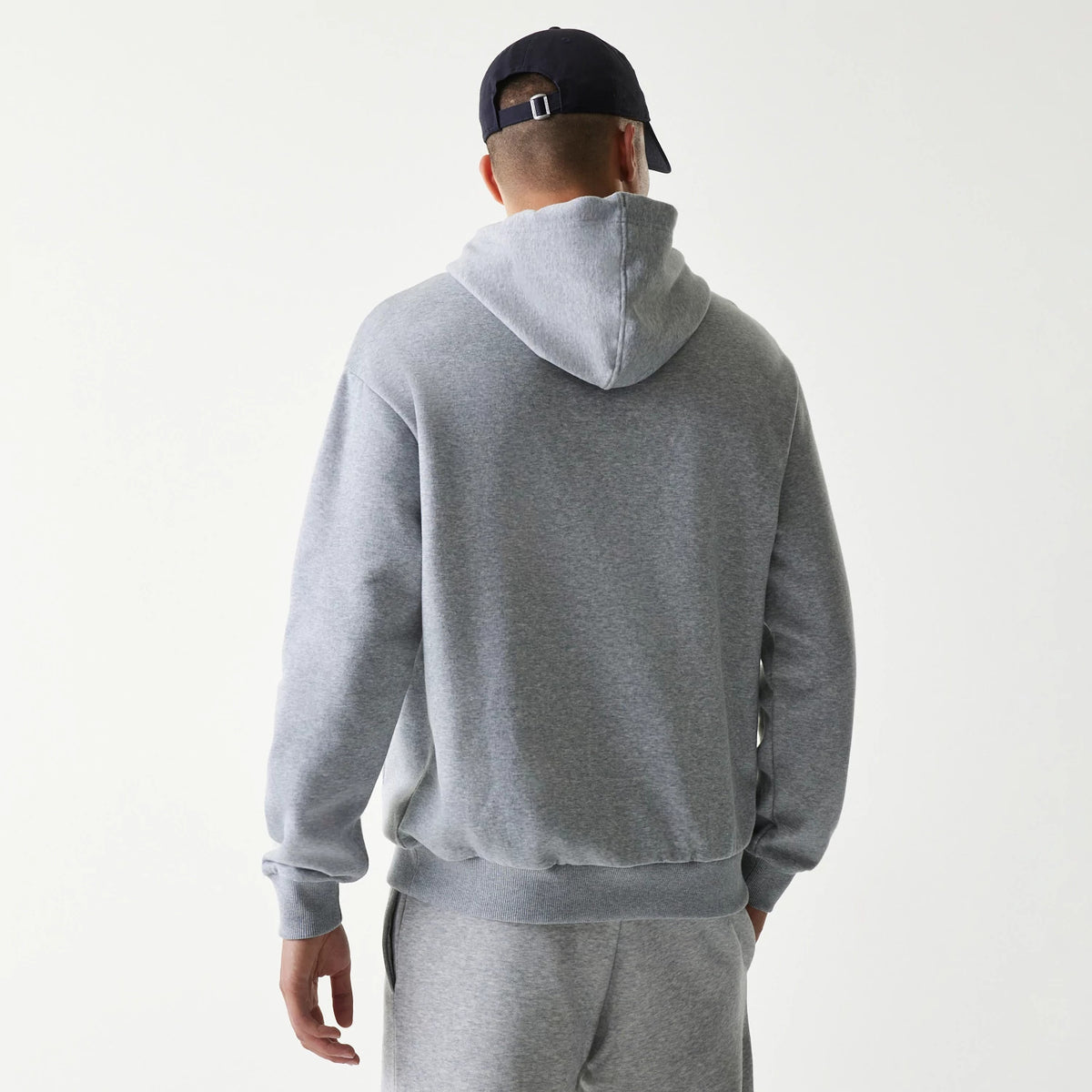 New Era New York Yankees MLB League Essential Grey Oversized Pullover Hoodie, back view showing the hood resting on the shoulders, ribbed cuffs, and hem, worn by a person in matching grey bottoms with a navy cap.
