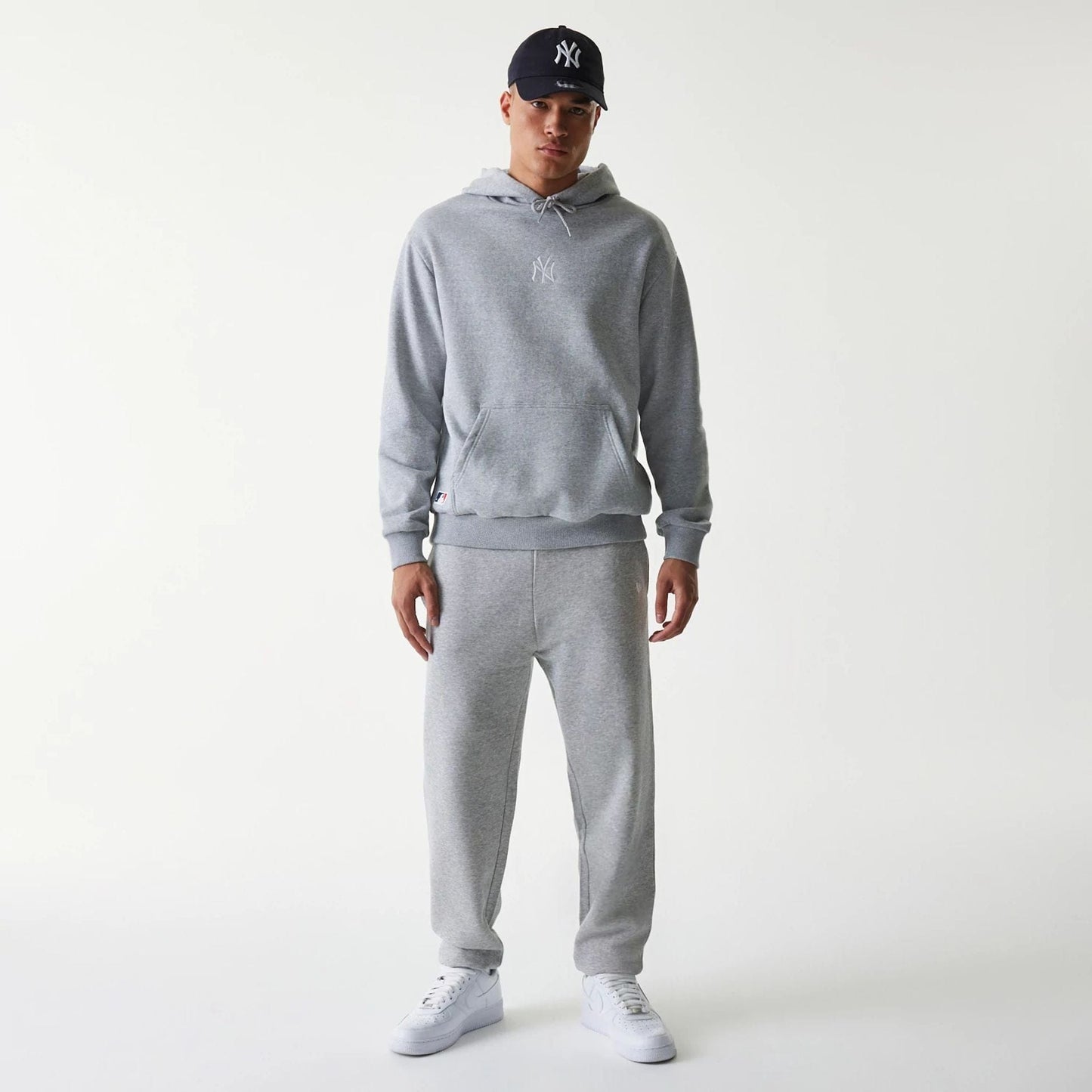 New Era New York Yankees MLB League Essential Grey Oversized Pullover Hoodie, on-model view showing the light grey hoodie with a kangaroo pocket and a white embroidered Yankees logo on the chest, paired with matching grey pants and white sneakers.