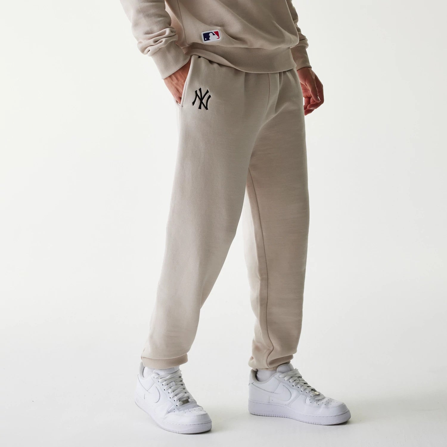 New Era New York Yankees MLB League Essential Cream Joggers, front view on a model wearing white trainers, featuring black NY logo embroidery on the left thigh and side pockets, with MLB logo on the hip of a matching sweatshirt.
