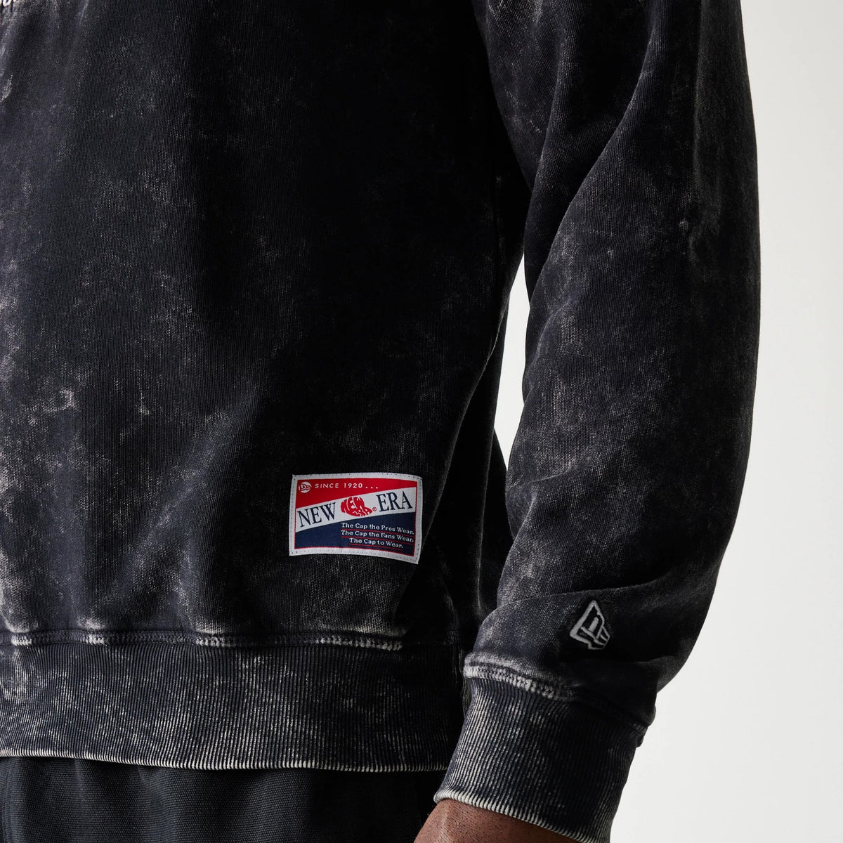 New Era Las Vegas Raiders NFL Throwback Black Crew Neck Sweater, close-up showing the New Era flag logo on the left sleeve and a vintage-style sewn label on the lower hem against a weathered black fabric.