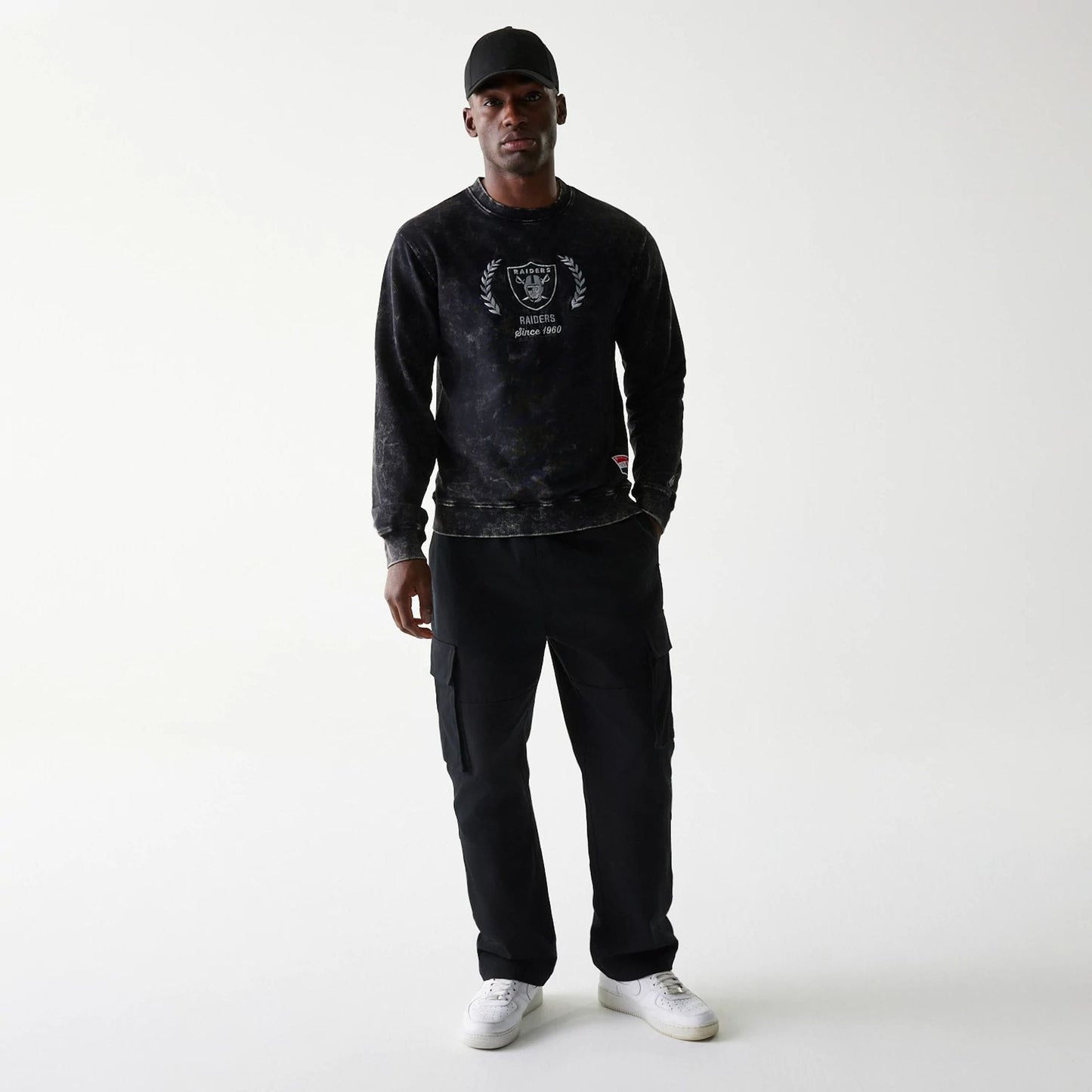 New Era Las Vegas Raiders NFL Throwback Black Crew Neck Sweater, on-model view with a black cap, featuring the Raiders logo with laurel leaves and Since 1960 text on the chest, paired with black cargo trousers and white trainers.