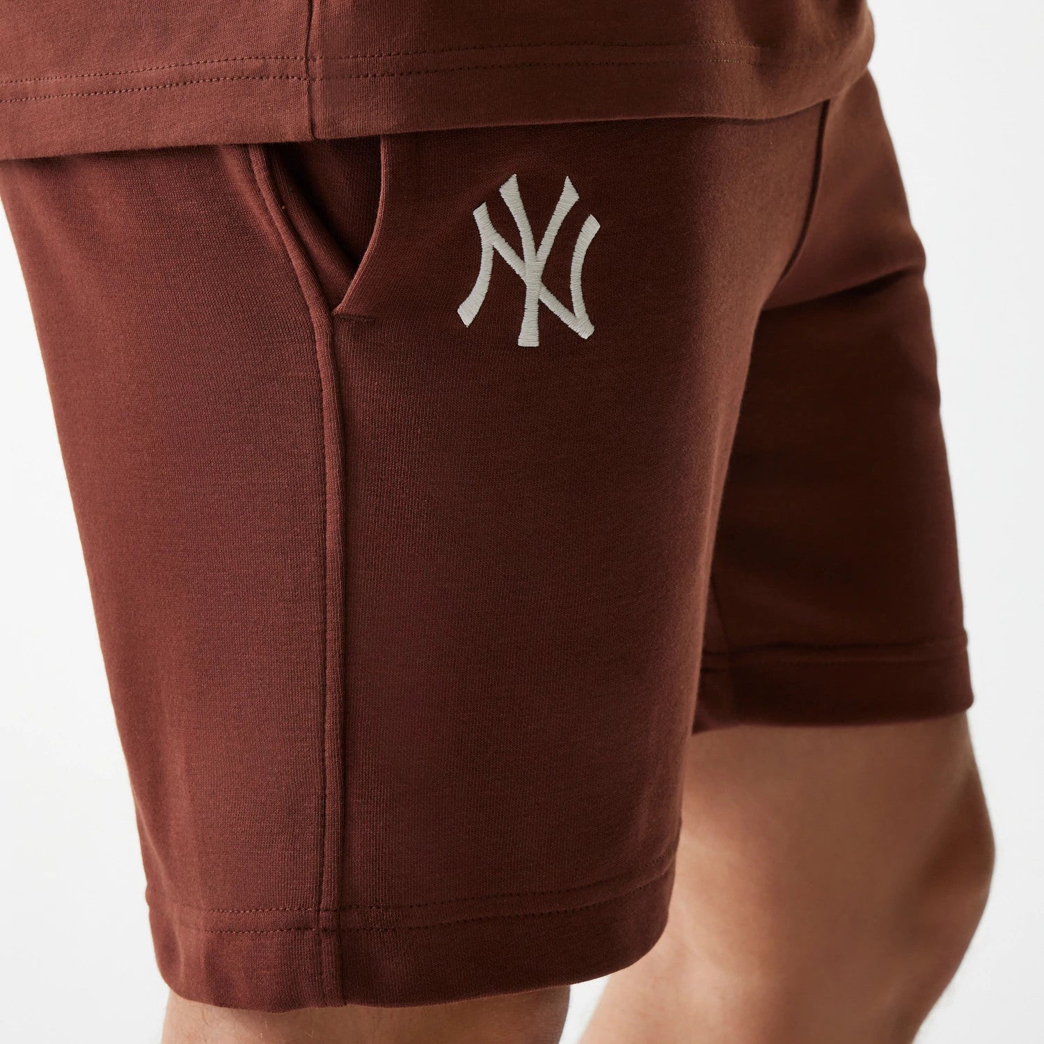 New Era New York Yankees MLB Midi League Essential Brown Shorts, front view showing the dark brown fabric with a white embroidered NY Yankees logo on the left thigh and a pocket seam running vertically beside it.