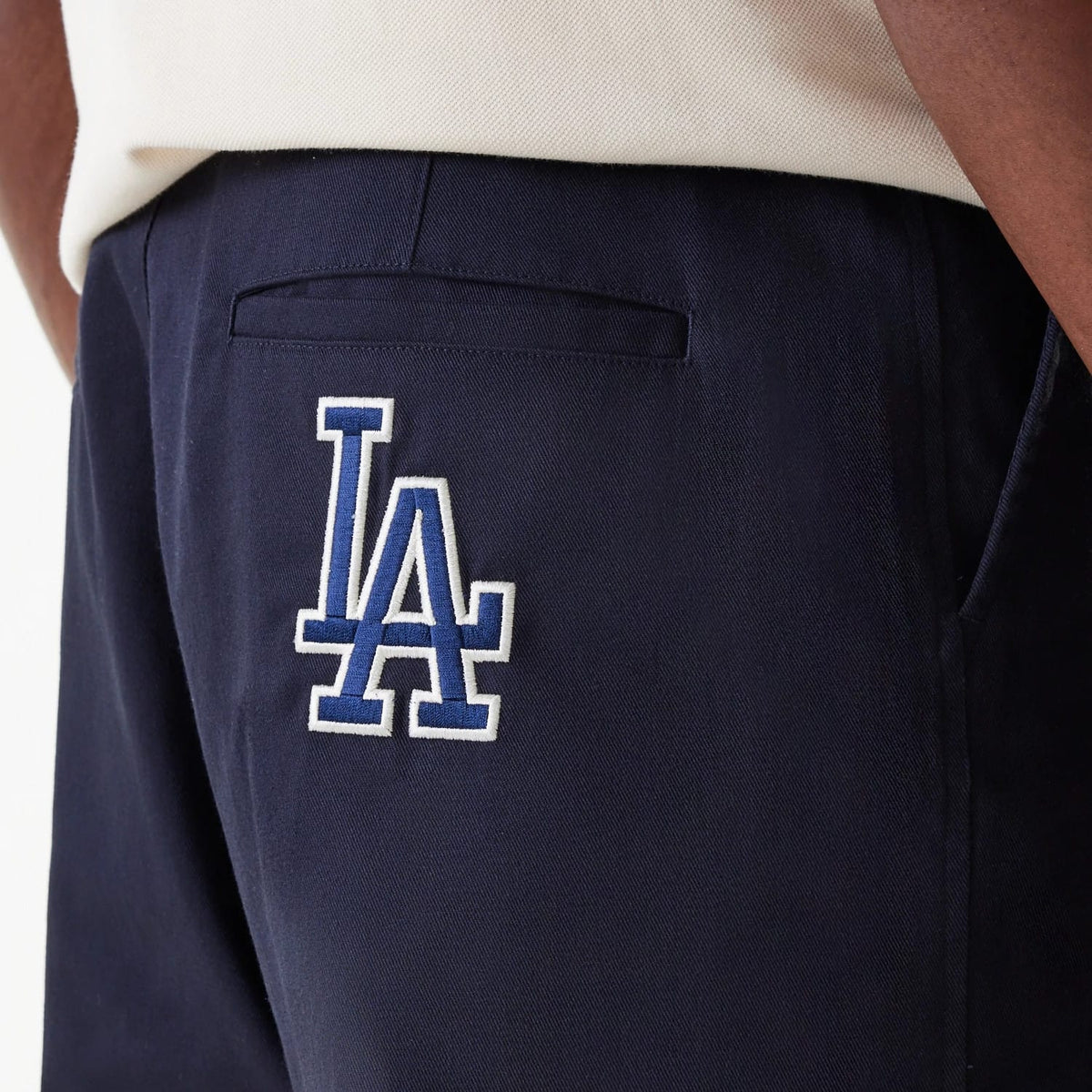 New Era LA Dodgers MLB Navy Chino Trousers, side view on a model showing the white and blue embroidered LA logo on the right back pocket below the waistband, contrasting with a cream shirt.